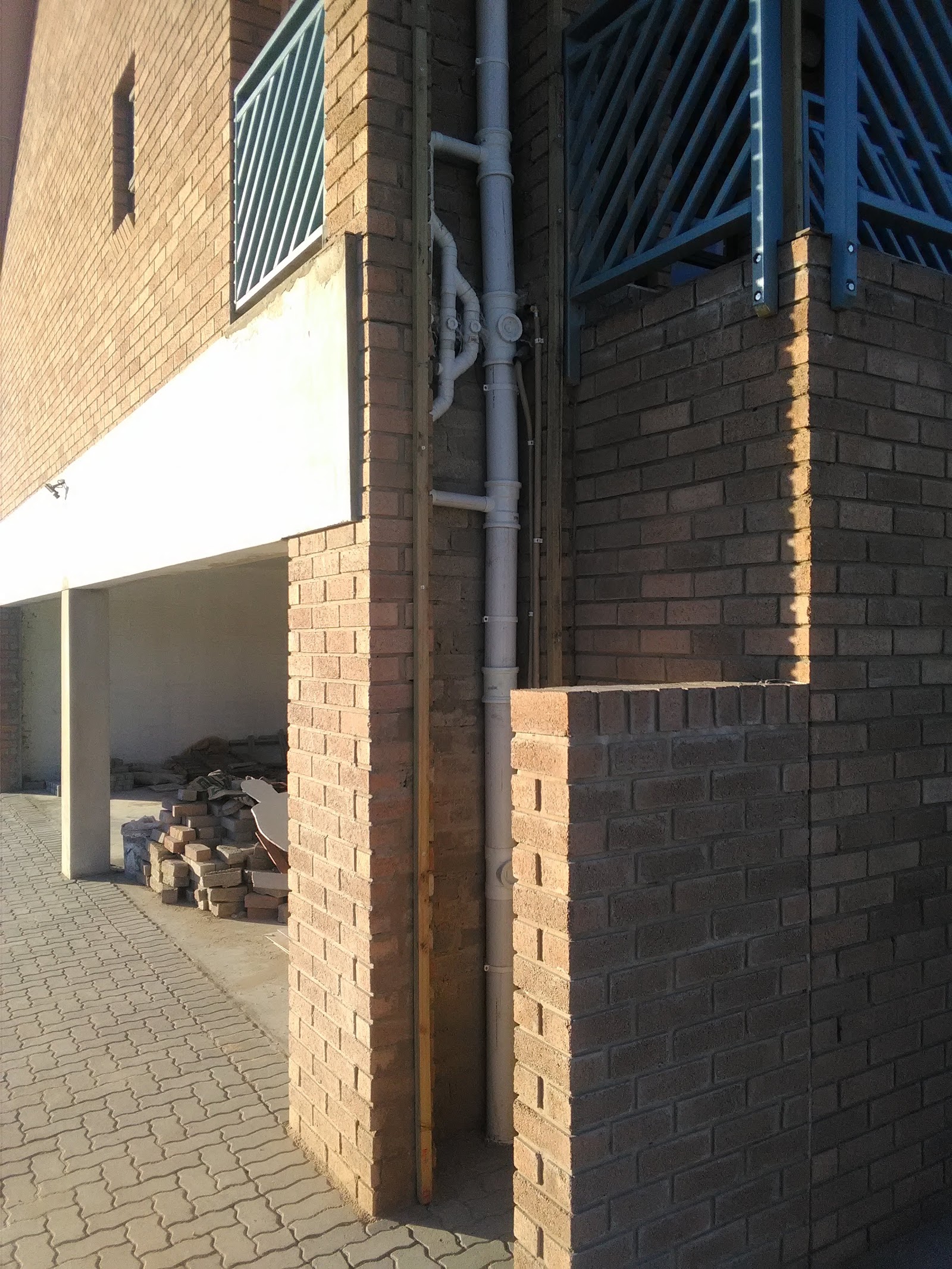Brick wall exterior with gray downpipe, metal railings, and stacked bricks in covered area.