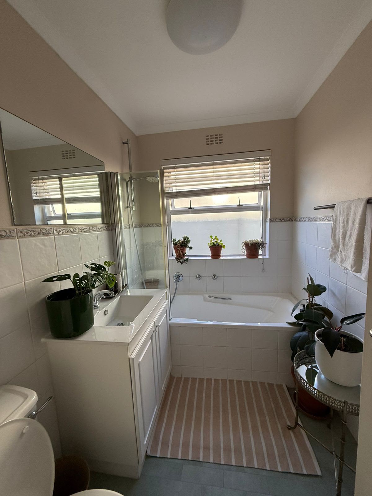 Bathroom with white bathtub, small window with plants, pedestal sink, and beige tile walls.