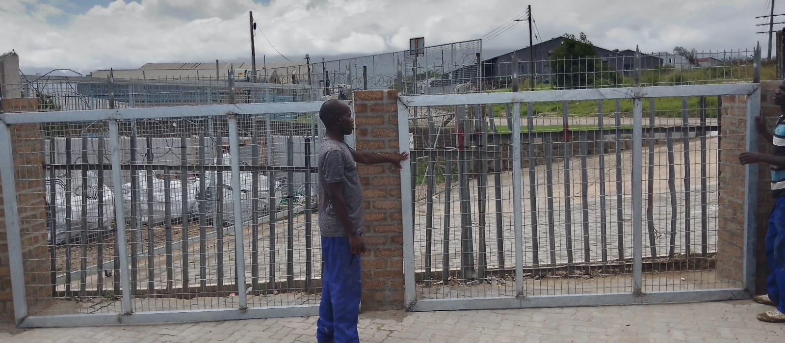 Worker in blue pants standing at metal gate entrance with brick pillars and fencing on industrial property.