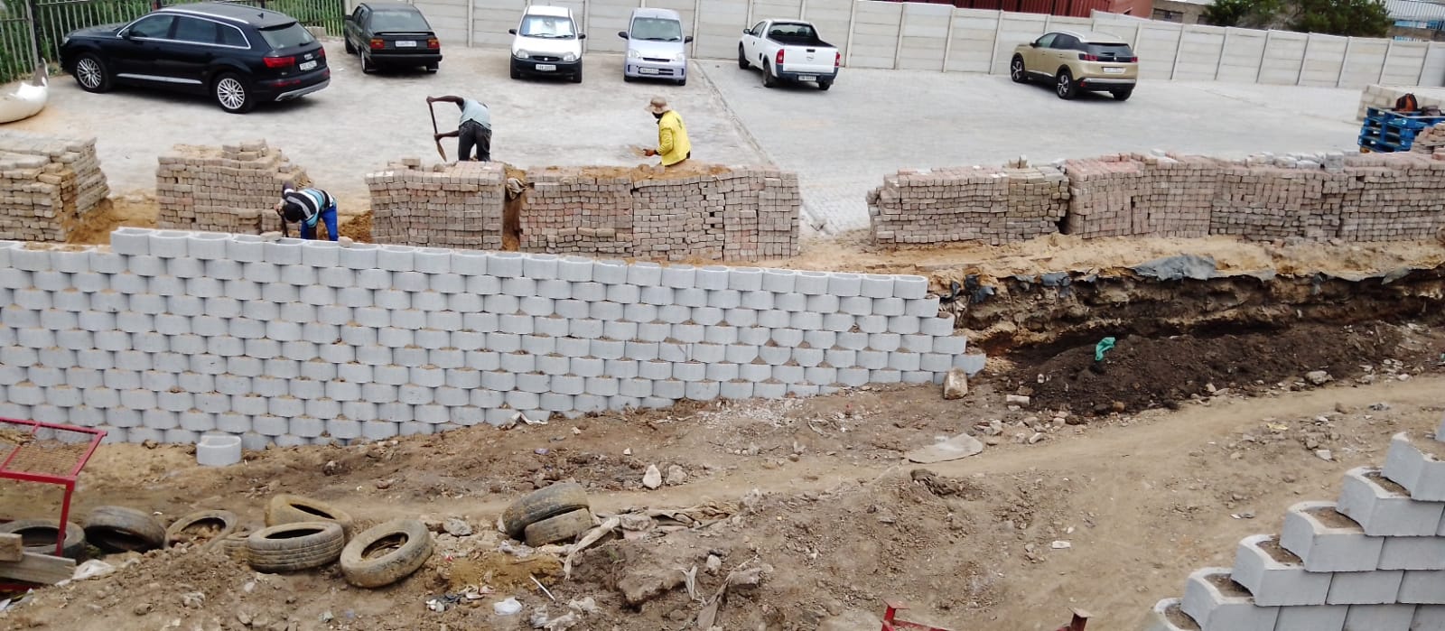 Multiple workers laying concrete blocks and red bricks with parked vehicles visible in secured yard.
