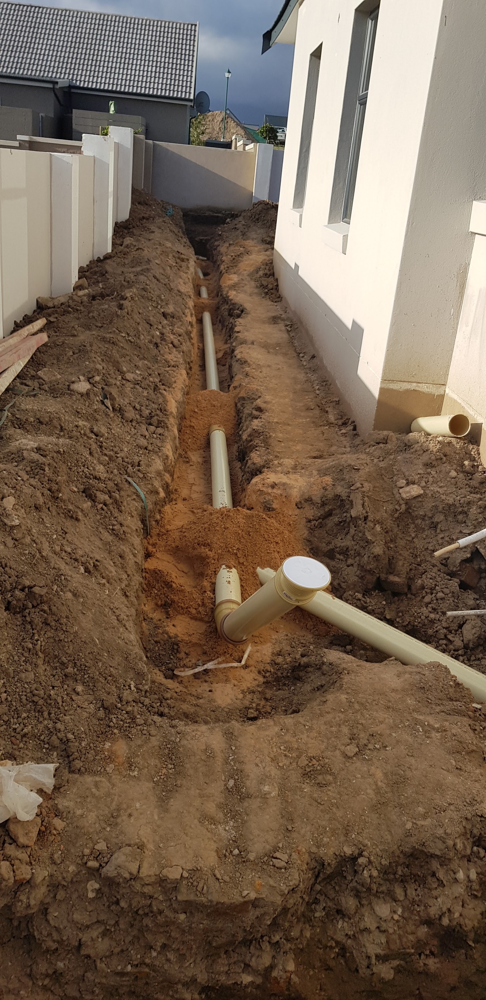 Excavated trench with PVC drainage pipes and fittings installed between house foundation and fence.
