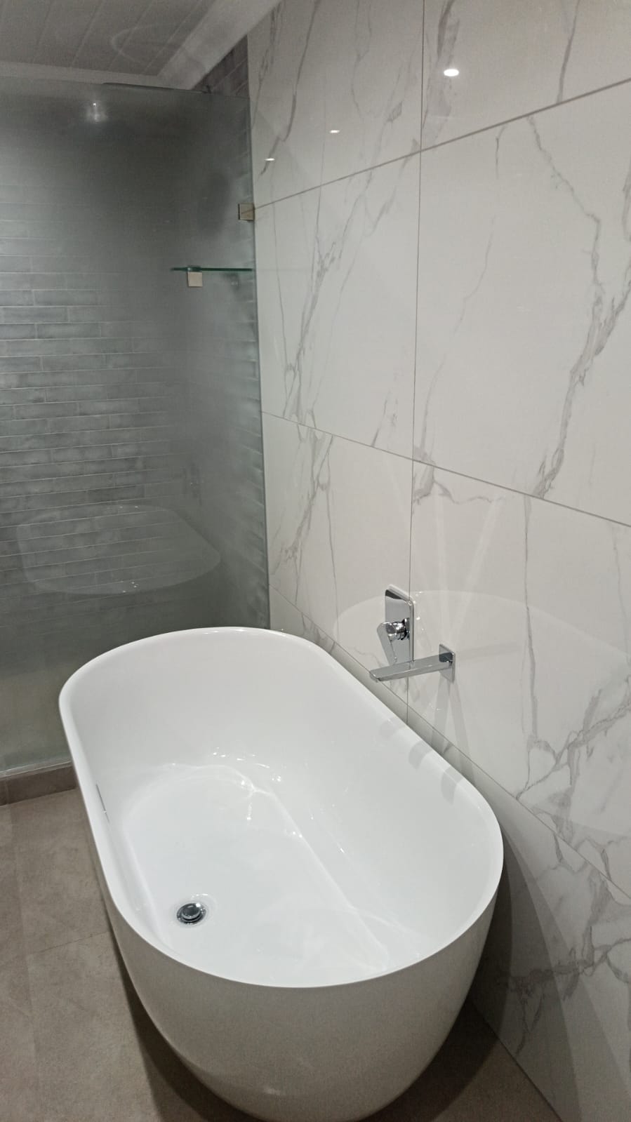 Modern bathroom featuring freestanding white tub, marble-look tile walls, and glass shower partition.