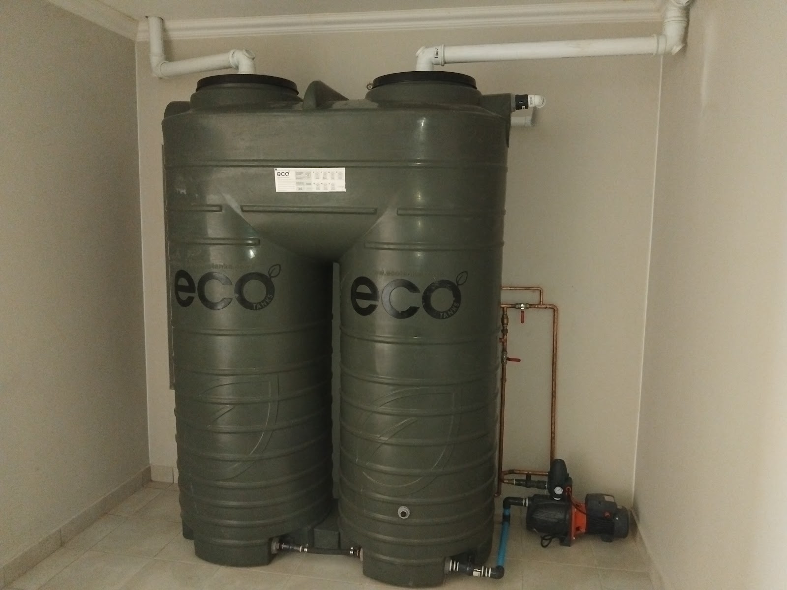 Two green Eco brand water tanks with connecting pipes and pump system installed indoors.