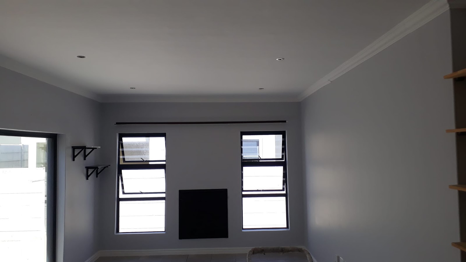 Empty room with recessed ceiling lights, black-framed windows, shelving brackets on wall.