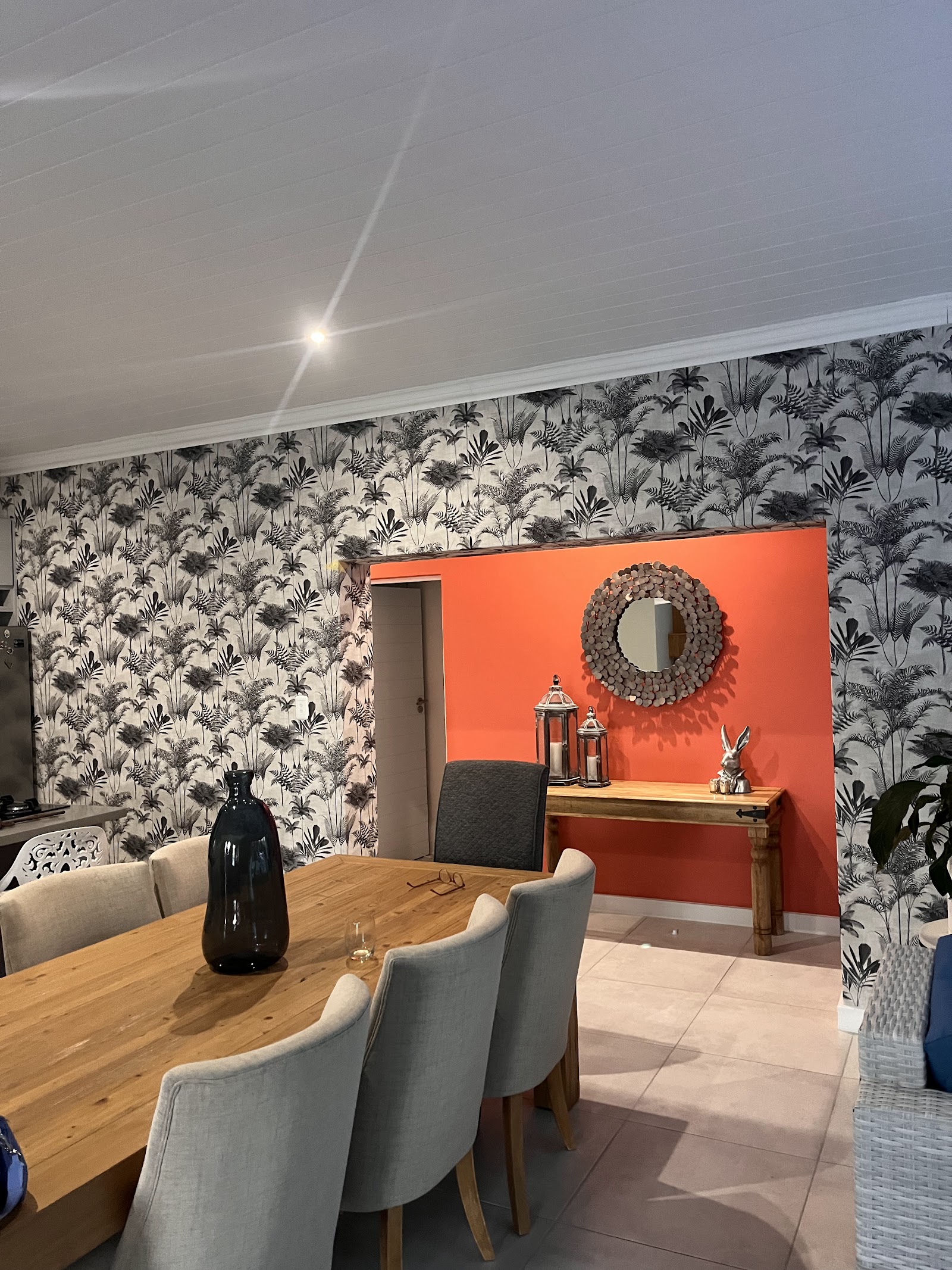 Interior showroom with tropical wallpaper, orange accent wall, wooden table, gray chairs, and decorative elements.