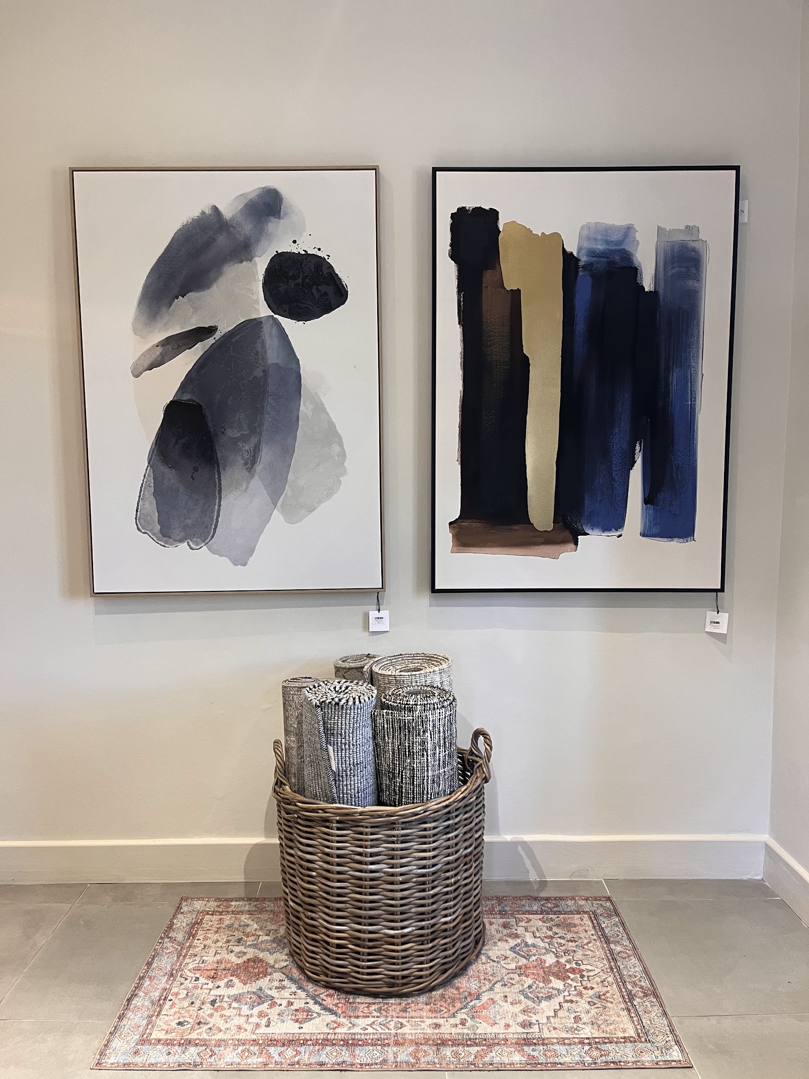 Two framed abstract watercolor artworks above wicker basket with rolled fabrics on patterned rug.