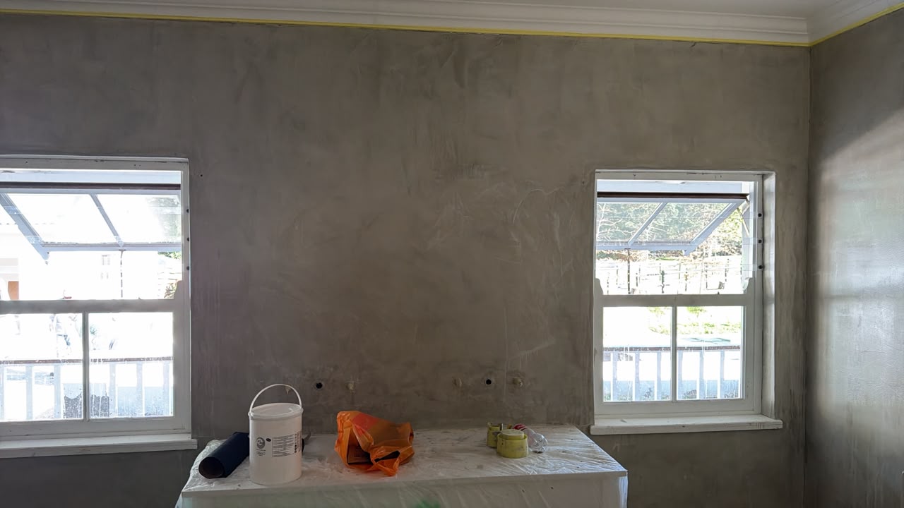 Drywall interior with two windows, paint bucket, orange tarp, and construction materials on table.