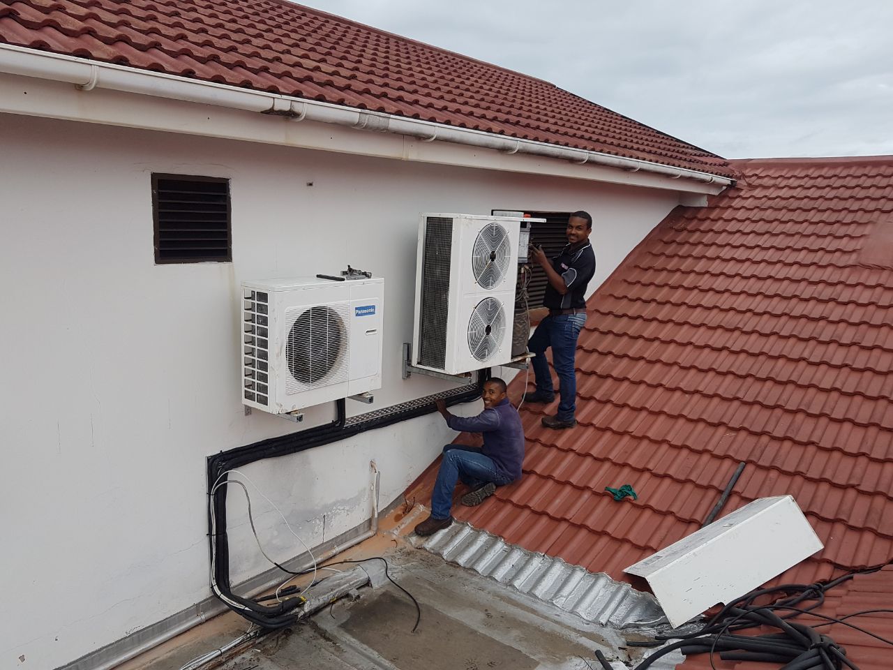 Two technicians installing Panasonic split air conditioning units on residential building exterior with red tile roof.