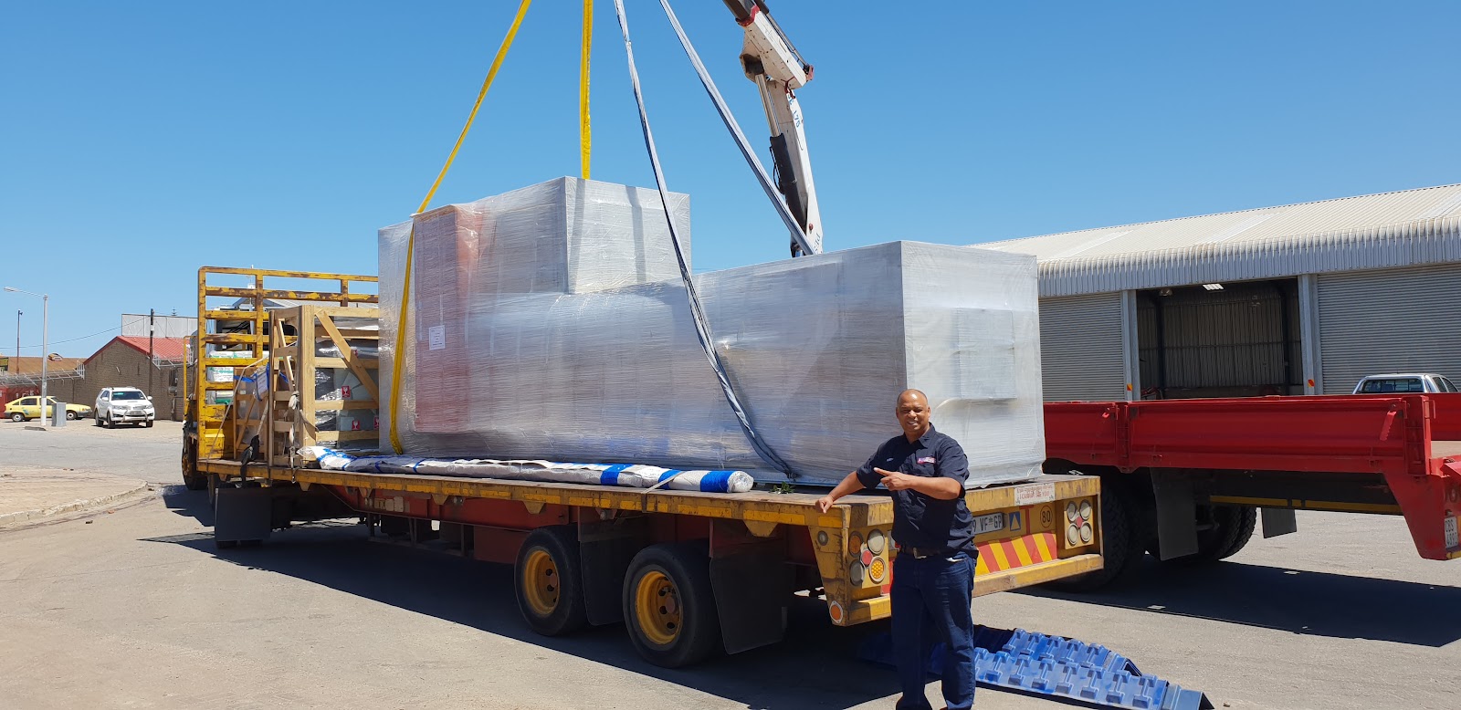Staff member with large wrapped commercial HVAC unit on flatbed truck with crane equipment at industrial yard.