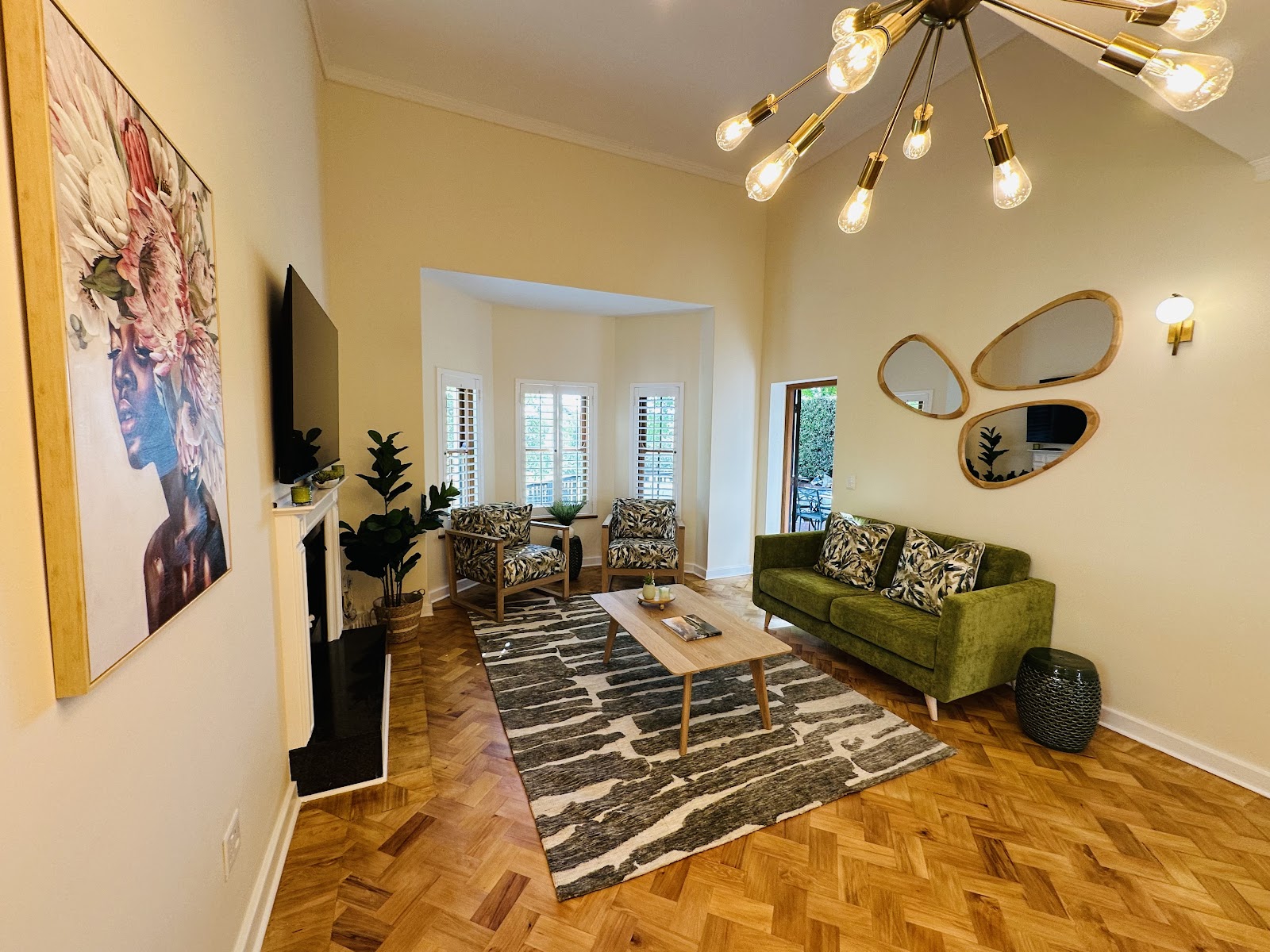 Modern living room with yellow walls, herringbone wood flooring, green sofa, brass pendant fixture, and decorative mirrors.