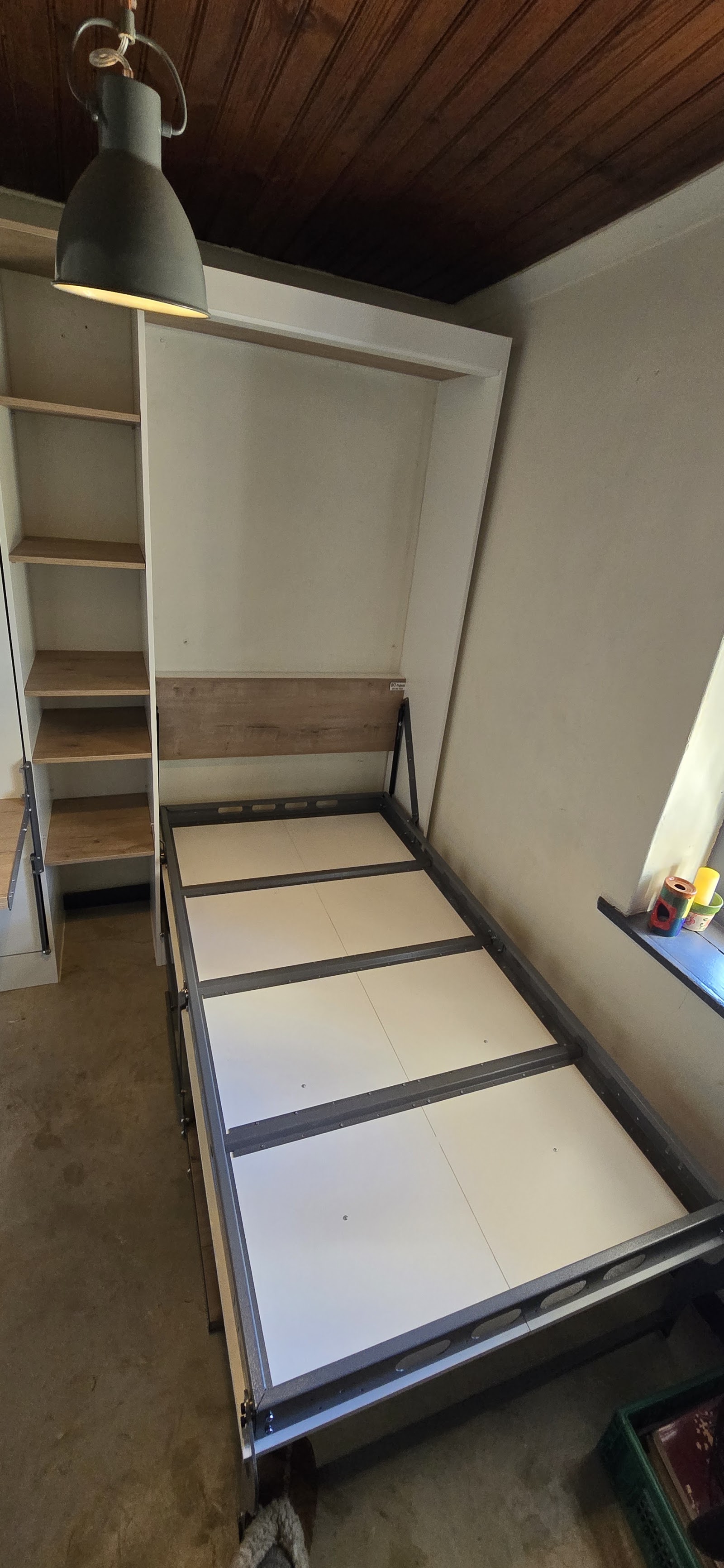 Bedroom interior featuring bed frame, built-in shelving, white walls, dark wood ceiling, and industrial pendant light.
