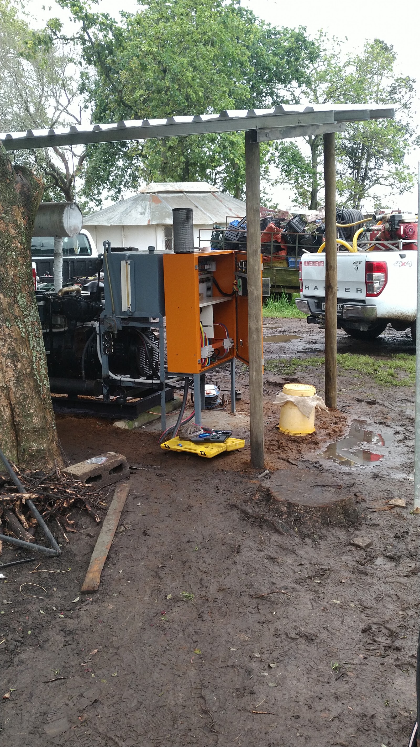 Orange diesel engine with fuel tank under pergola, surrounded by industrial equipment and vehicles on muddy lot.