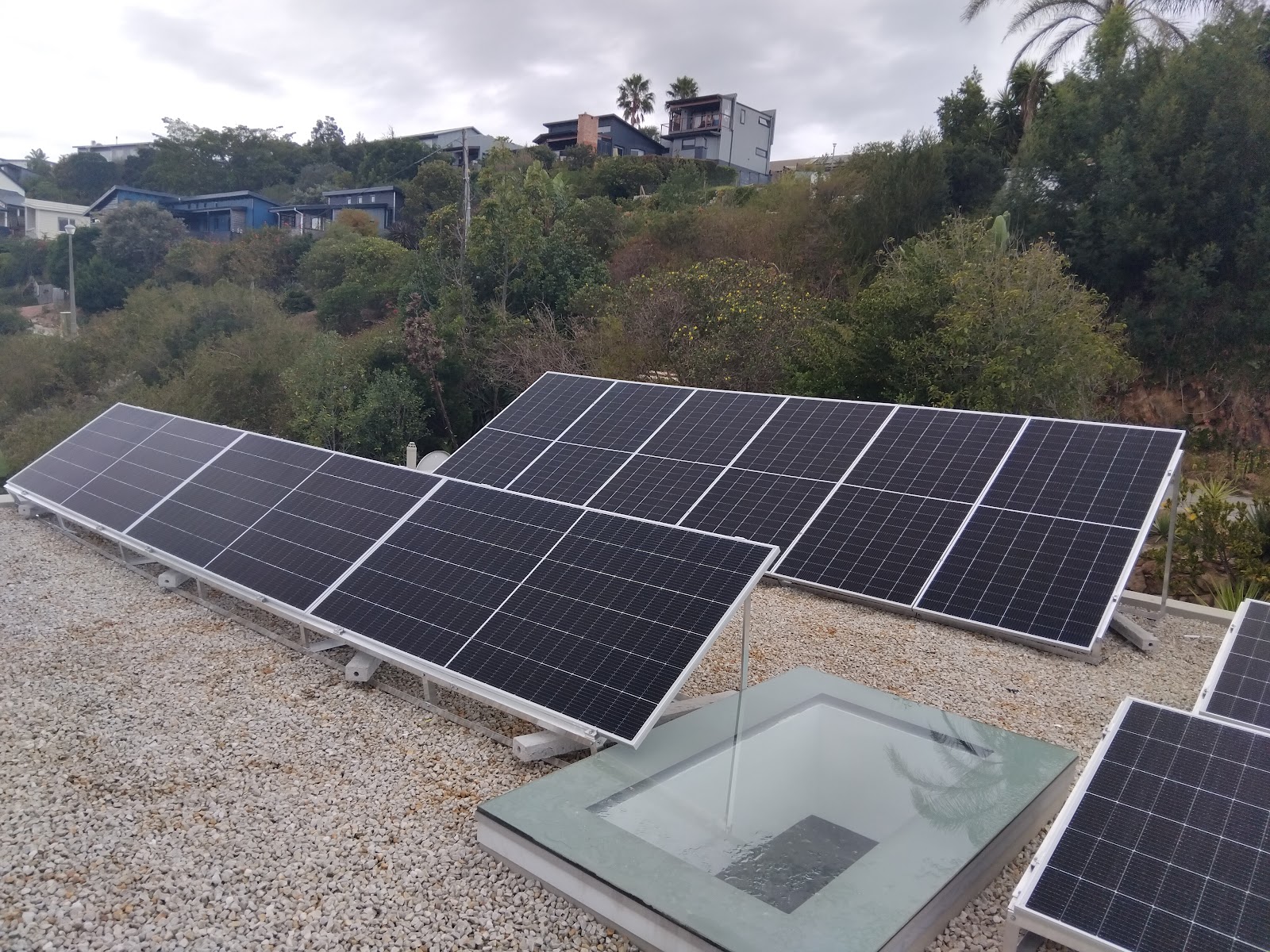 Solar panel array installation on ground-mounted gravel base with hillside residential buildings visible.