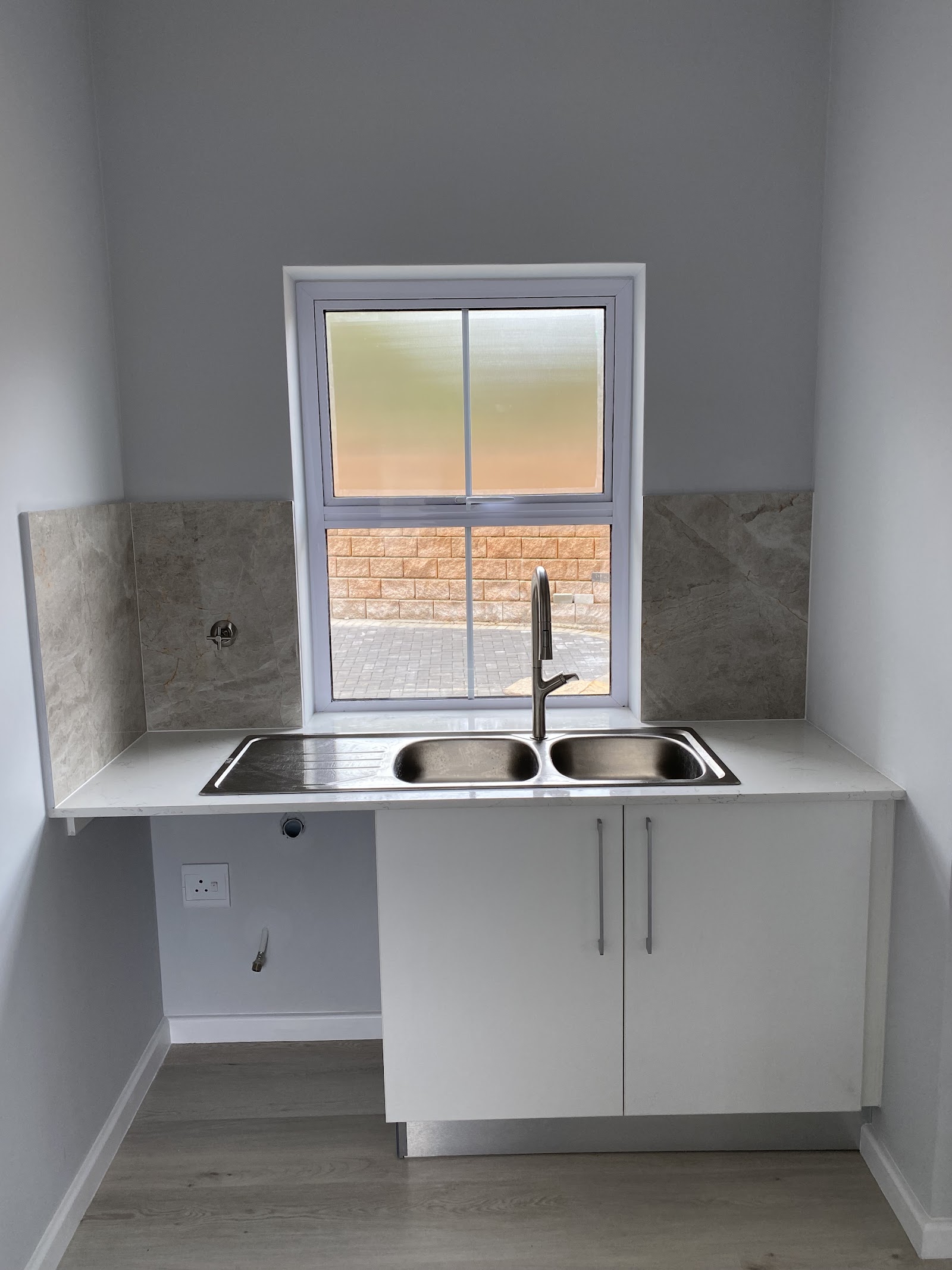 Compact kitchenette with white cabinetry, double stainless steel sink, gray tile backsplash, and window view.