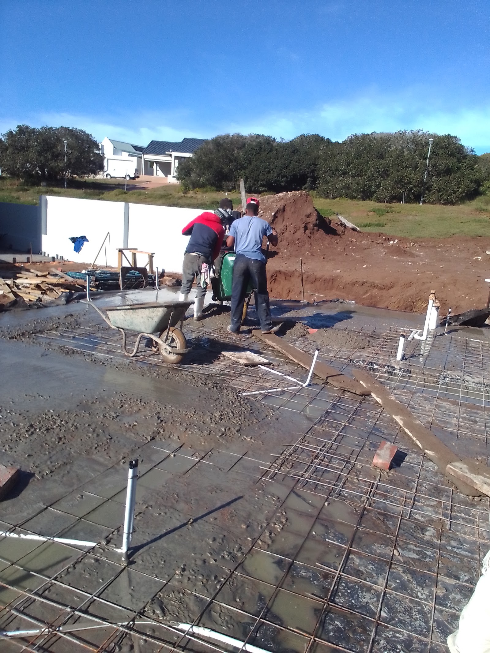 Two workers pouring concrete foundation with steel reinforcement mesh and wheelbarrow on residential site.