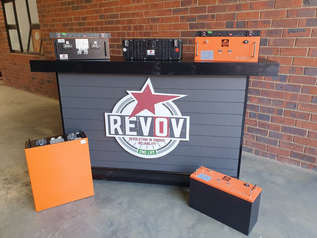 REVOV energy storage system display with orange and black battery units and branded signage against brick wall.