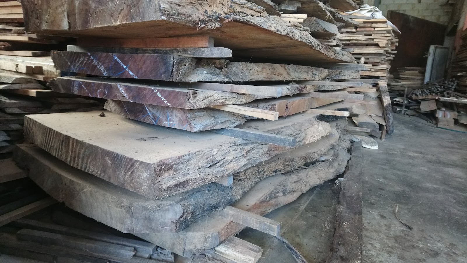 Mixed lumber and rough-cut timber pieces stacked indoors with plastic sheeting covers.