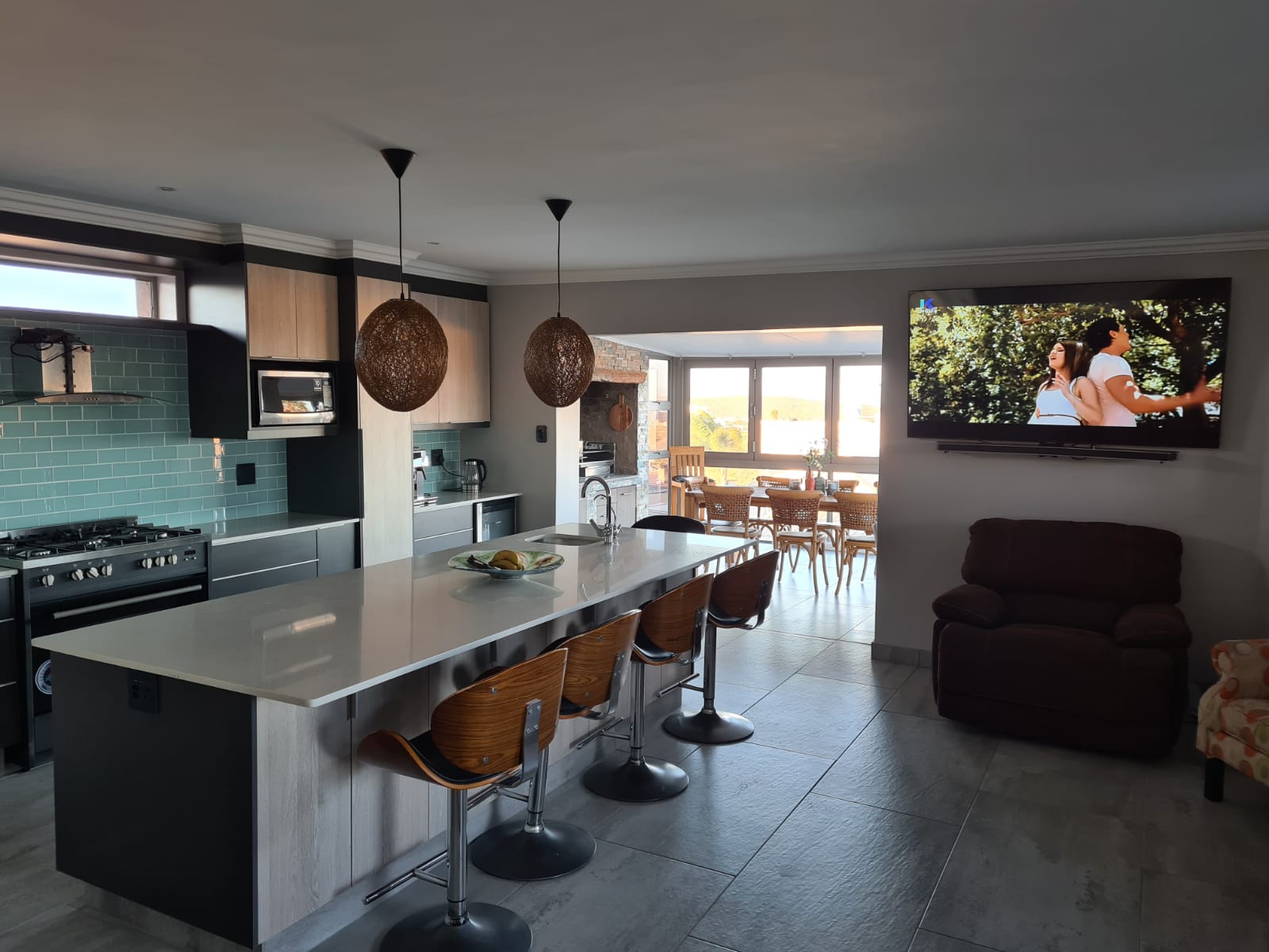 Modern kitchen-living space with woven pendant lights, island seating, turquoise tile backsplash, ocean views.