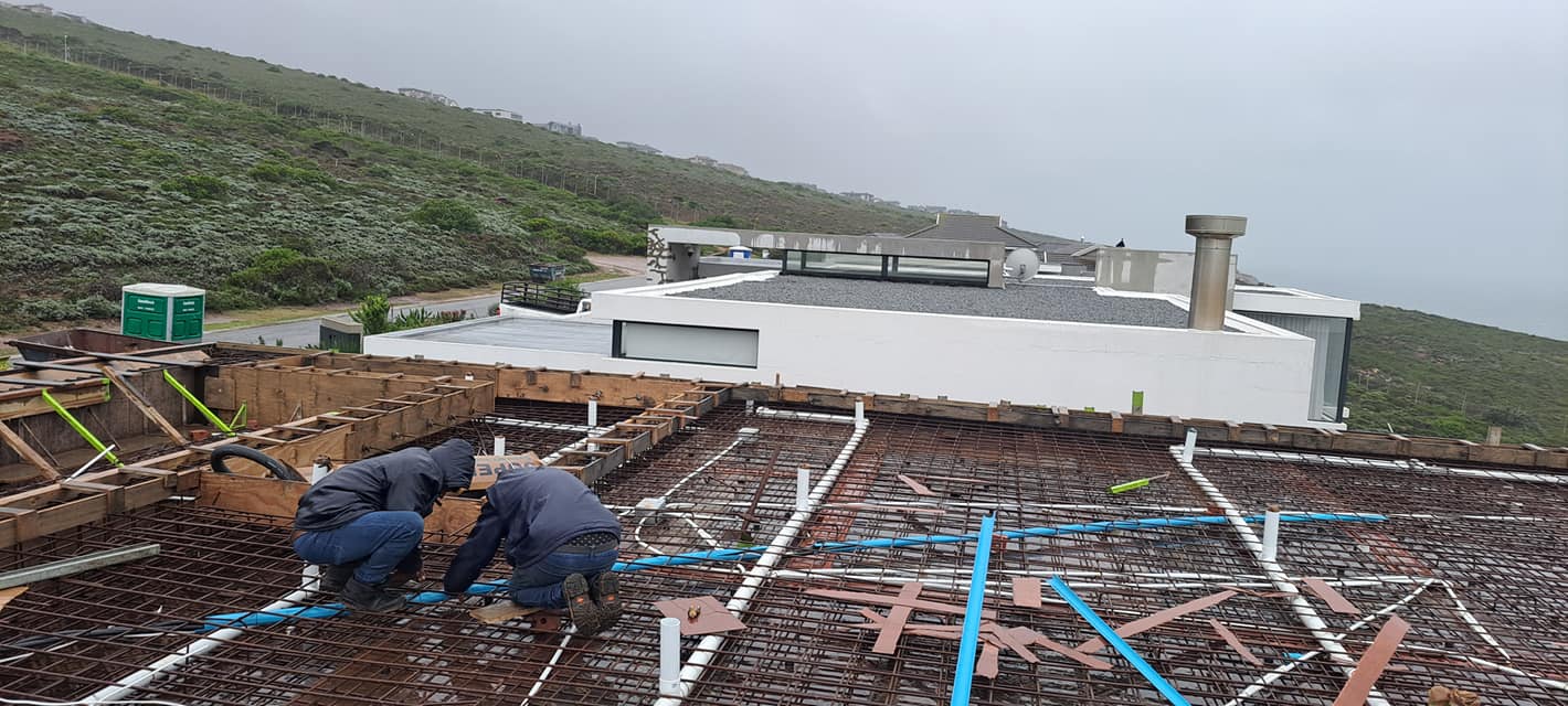 Two technicians in navy uniforms installing underfloor heating system with blue piping on residential foundation.