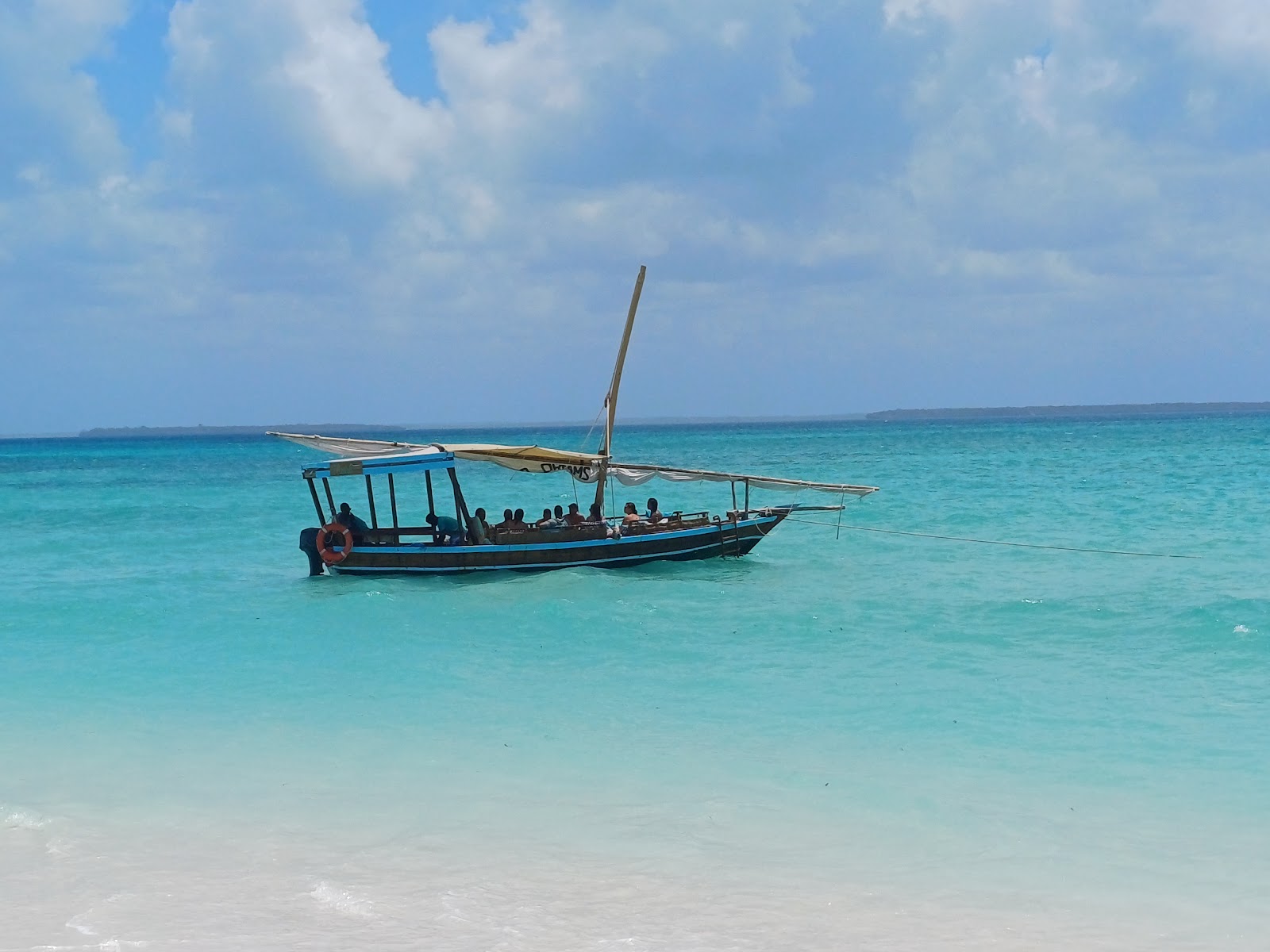Traditional wooden dhow boat with multiple passengers in turquoise tropical waters and beach.