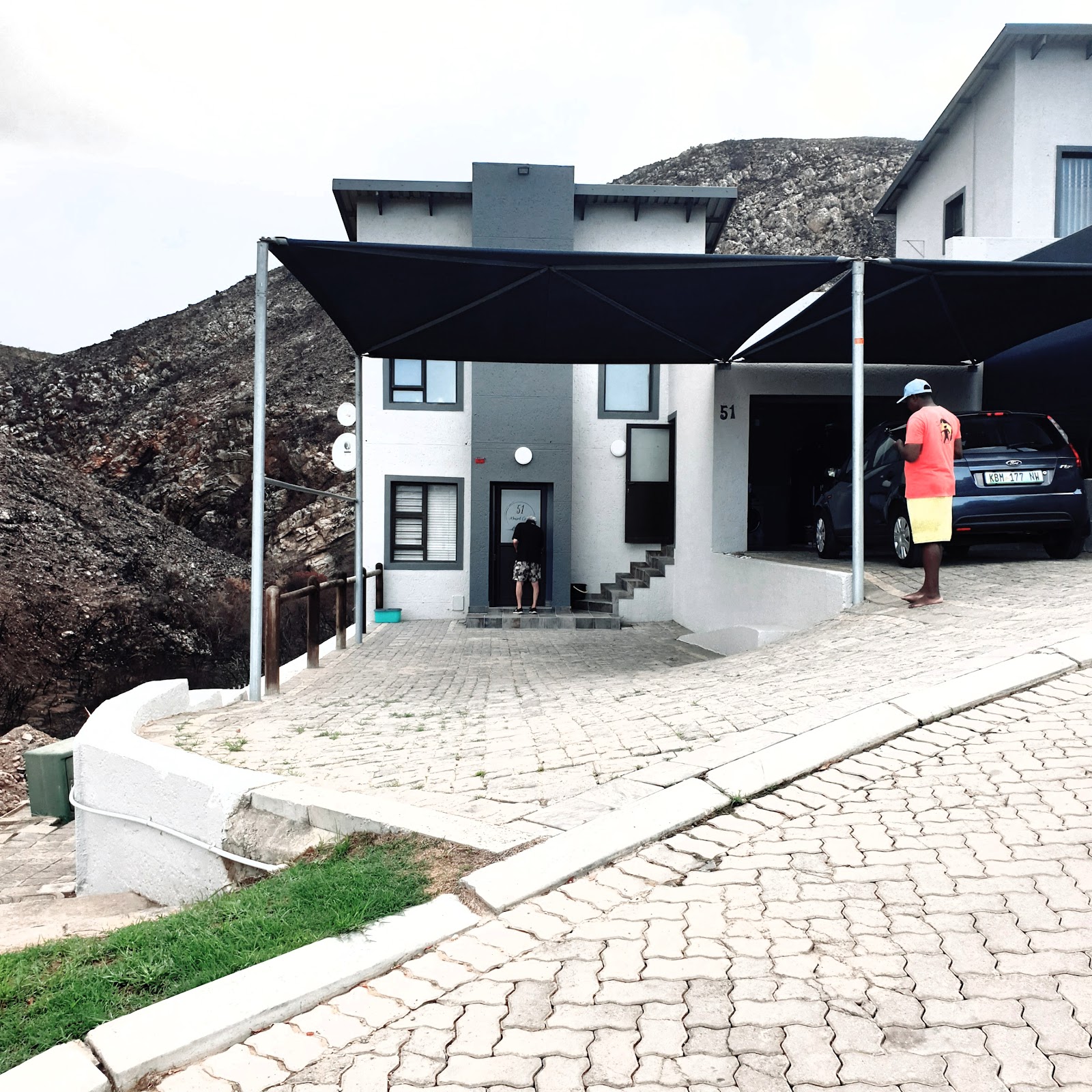 Staff member in safety vest directing parked car into carport beside white modern house numbered 51 with black canopy.
