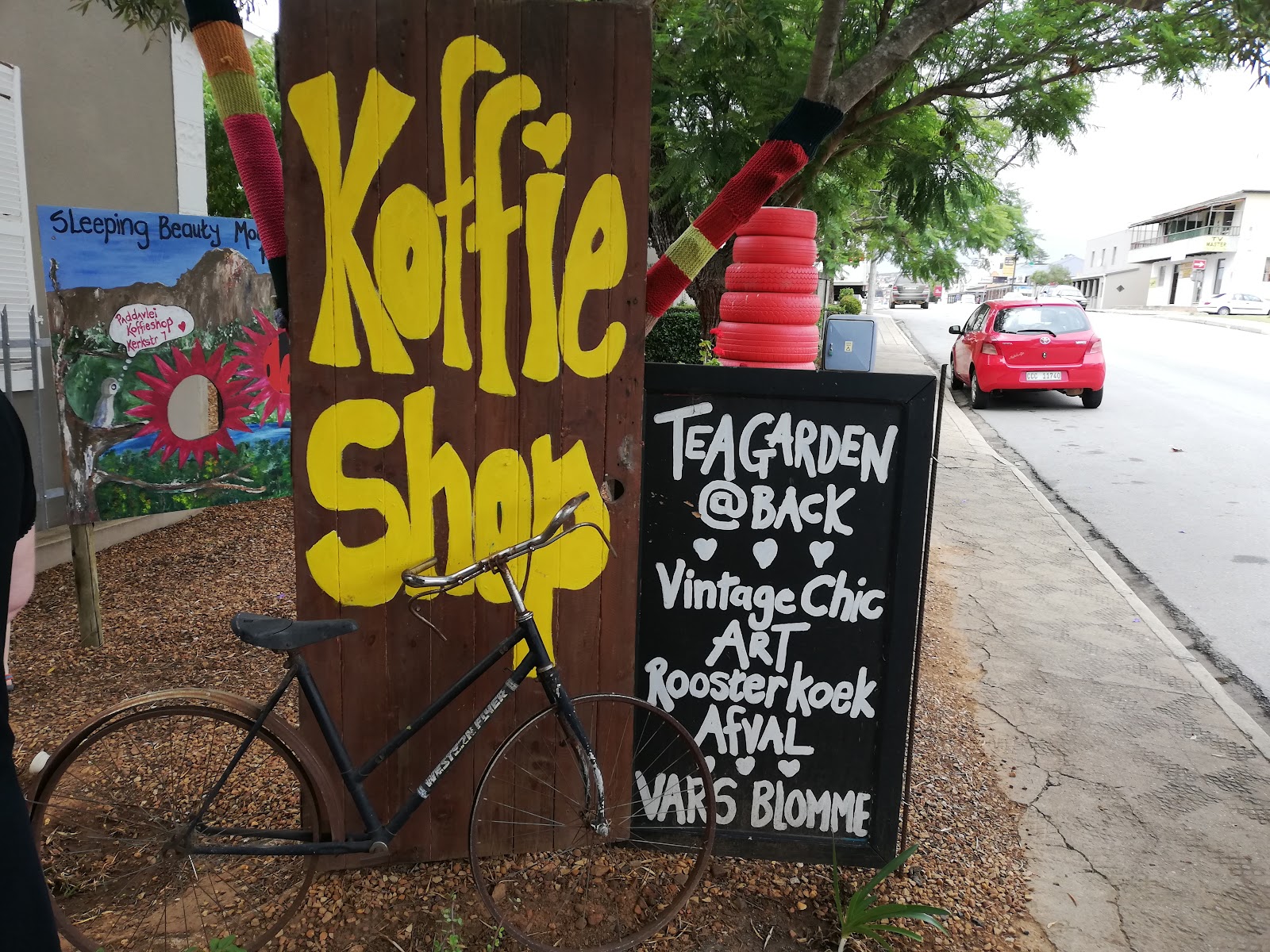 "Koffie Shop" storefront with yellow lettering, tea garden signage, bicycle, and street-facing wooden sign display.
