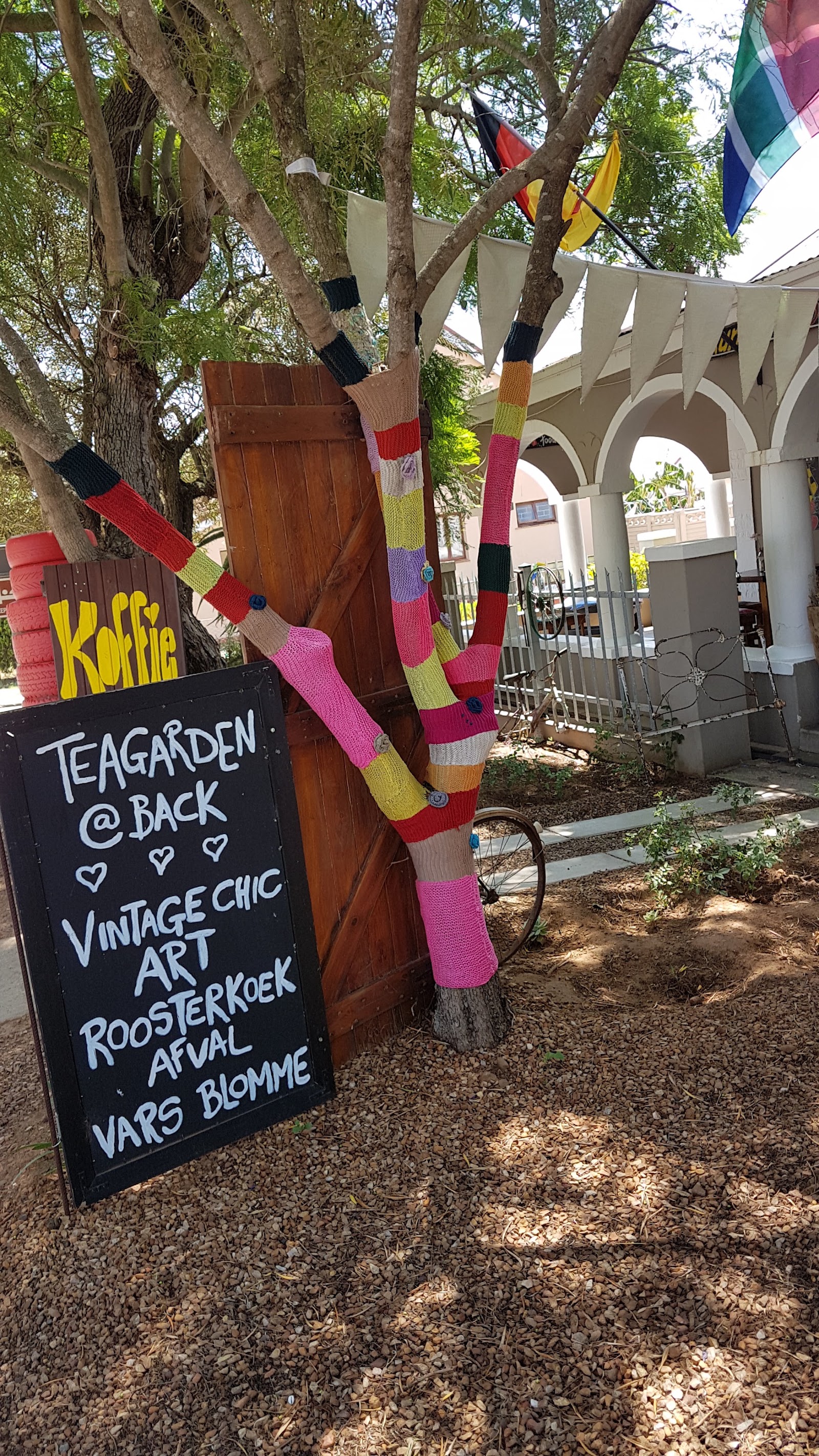 "Koffie" sign with hand-knitted tree wrapping, tea garden signage, colorful flags, and white pergola structure.
