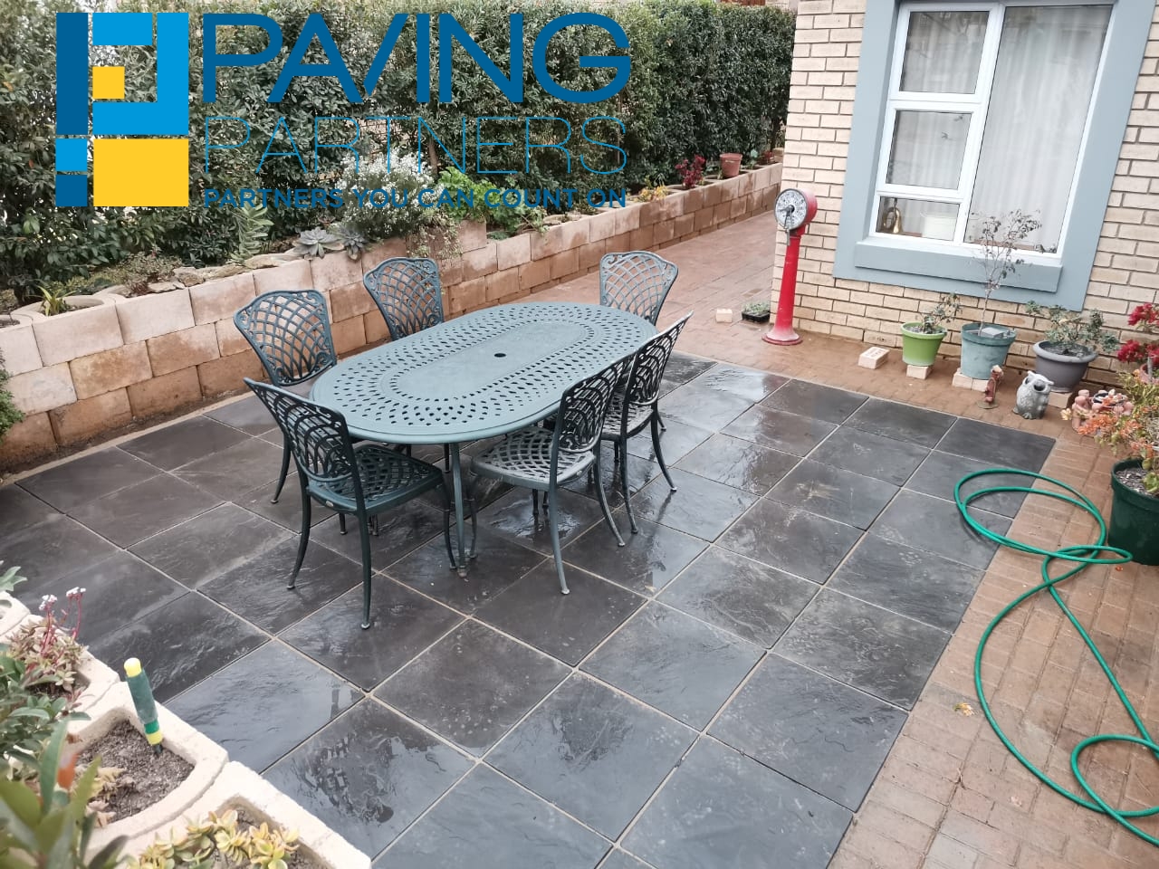 Paving Partners branding on residential slate patio with outdoor dining set and brick retaining wall.