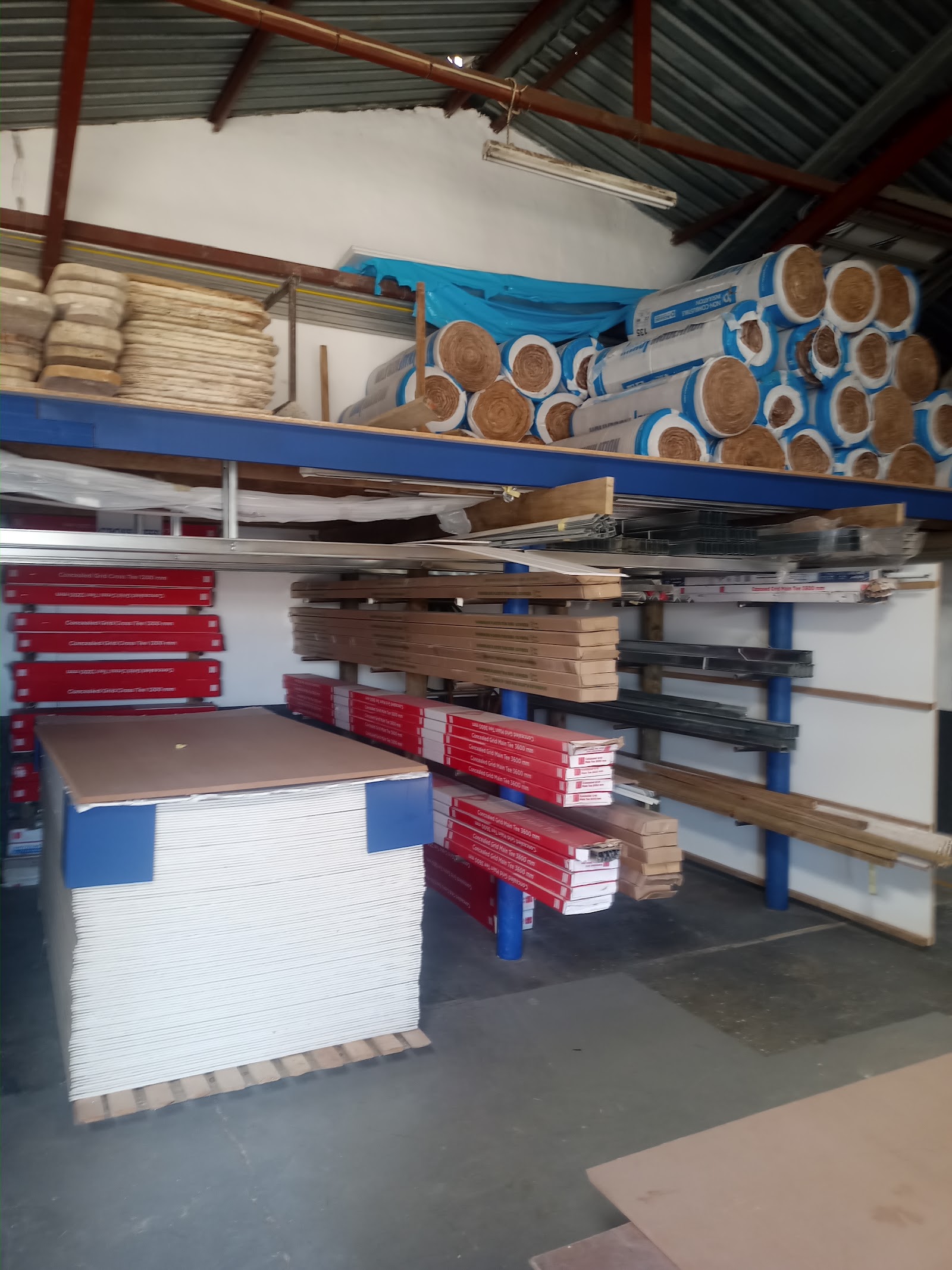Warehouse storage with blue racking system holding insulation rolls, drywall sheets, and building materials.