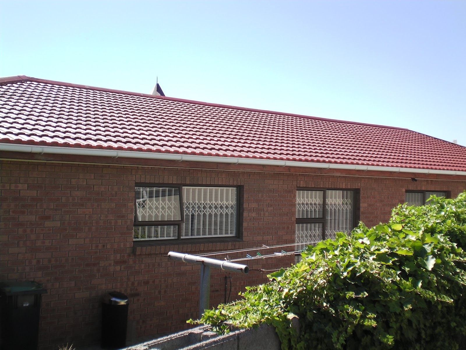 Completed red tile roof on single-story brick residential building with security bars and landscaping.