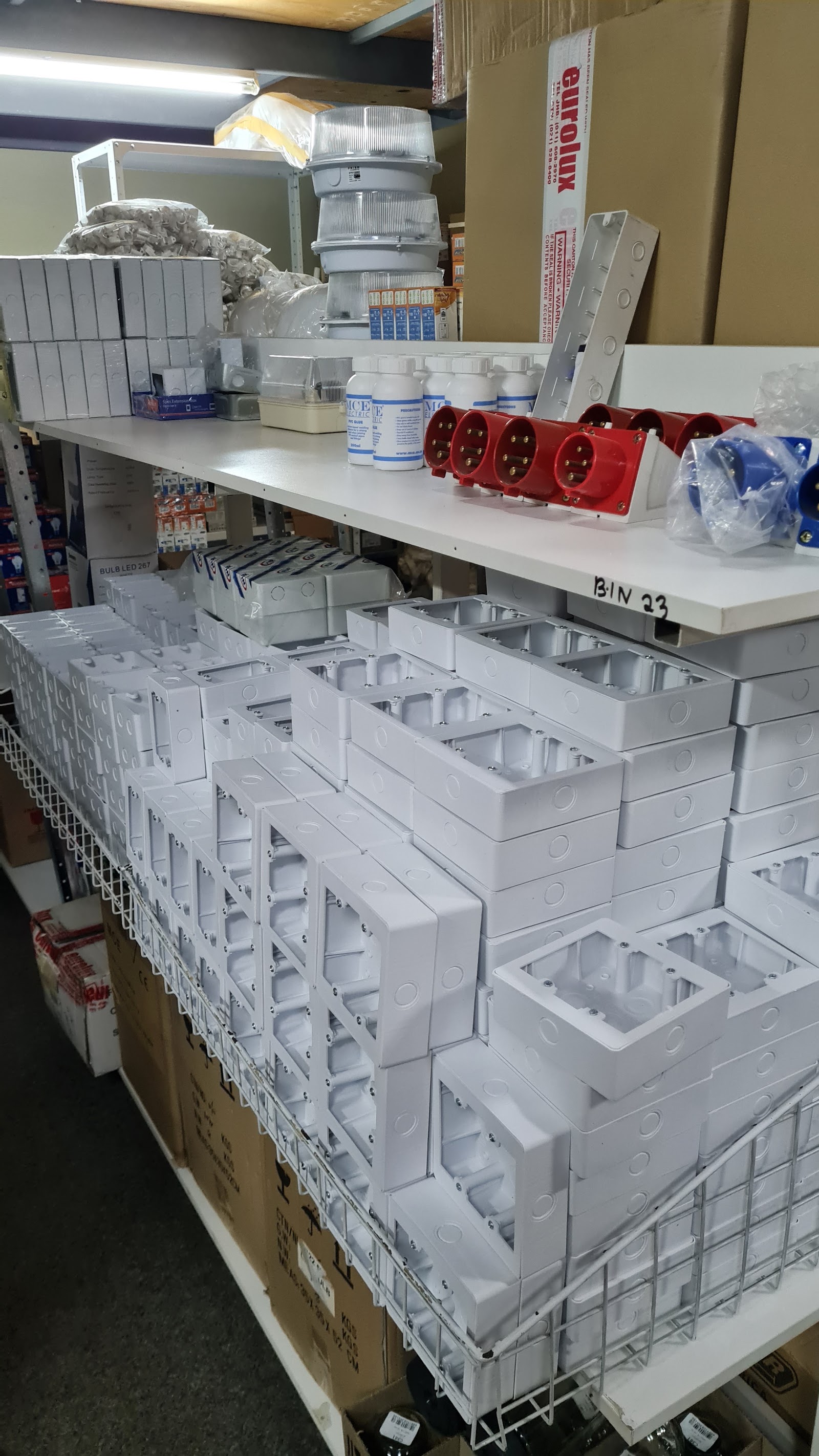Storage shelves with white electrical junction boxes, red power outlets, and miscellaneous wiring components in bins.