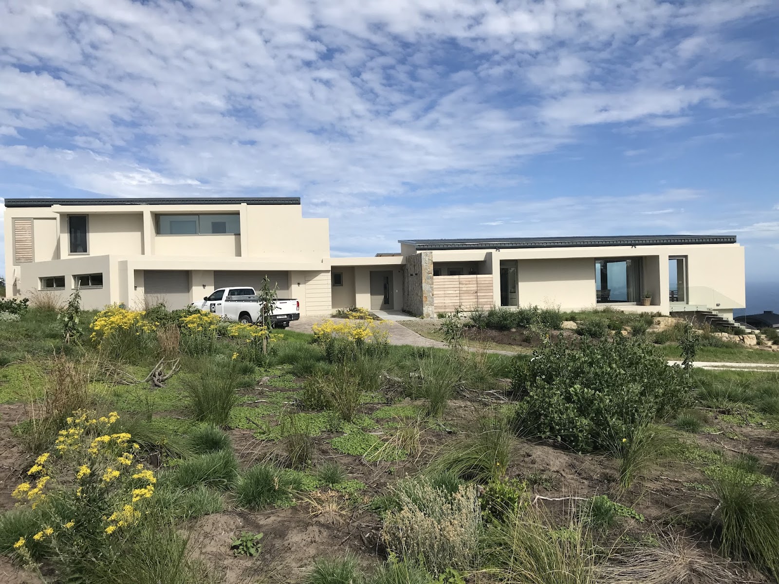 Completed contemporary residential home with cream render, black roof trim, white pickup truck, and native plantings.
