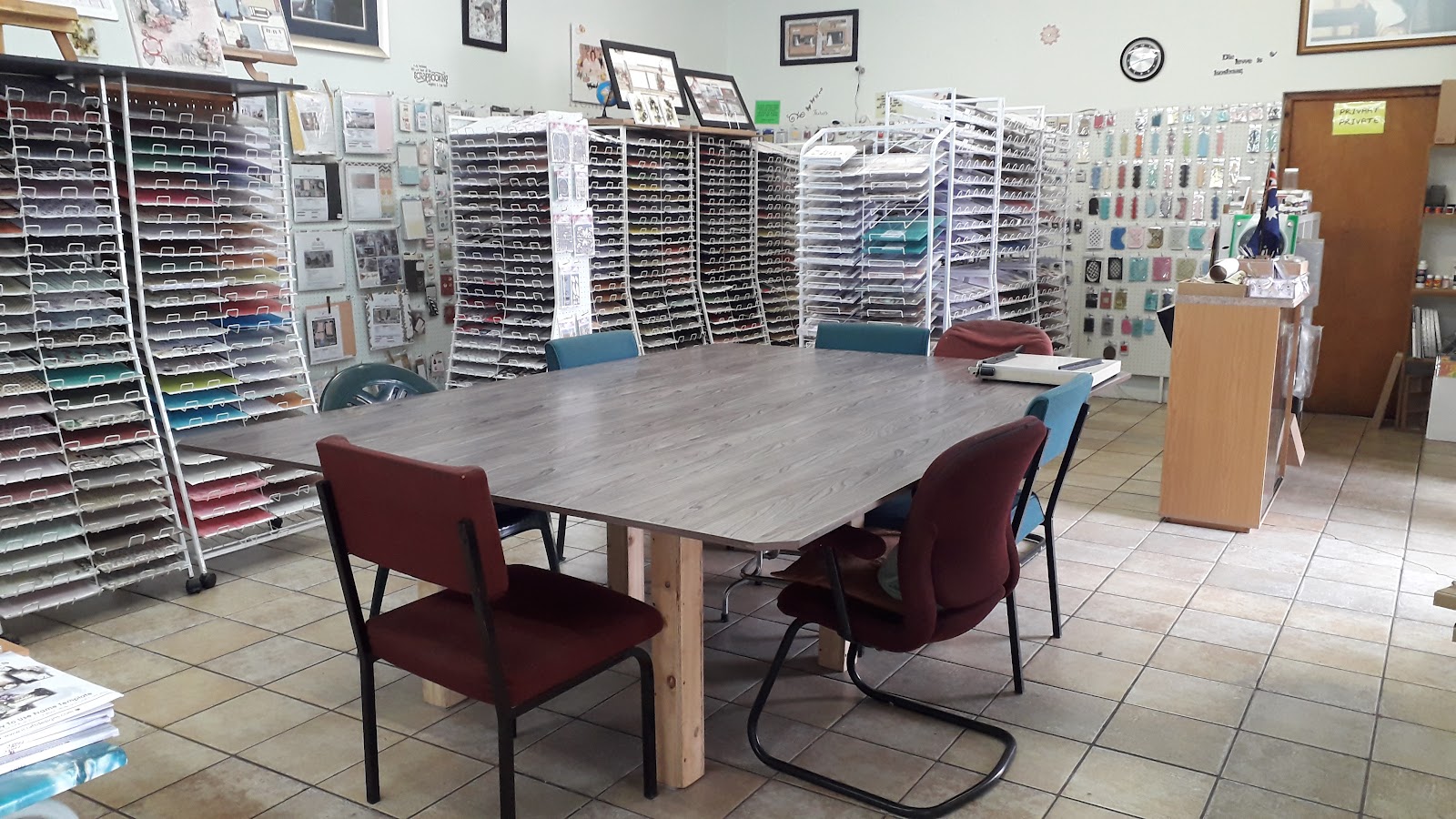 Interior craft workspace with organized shelving units displaying papers, embellishments, workspace table with burgundy chairs.