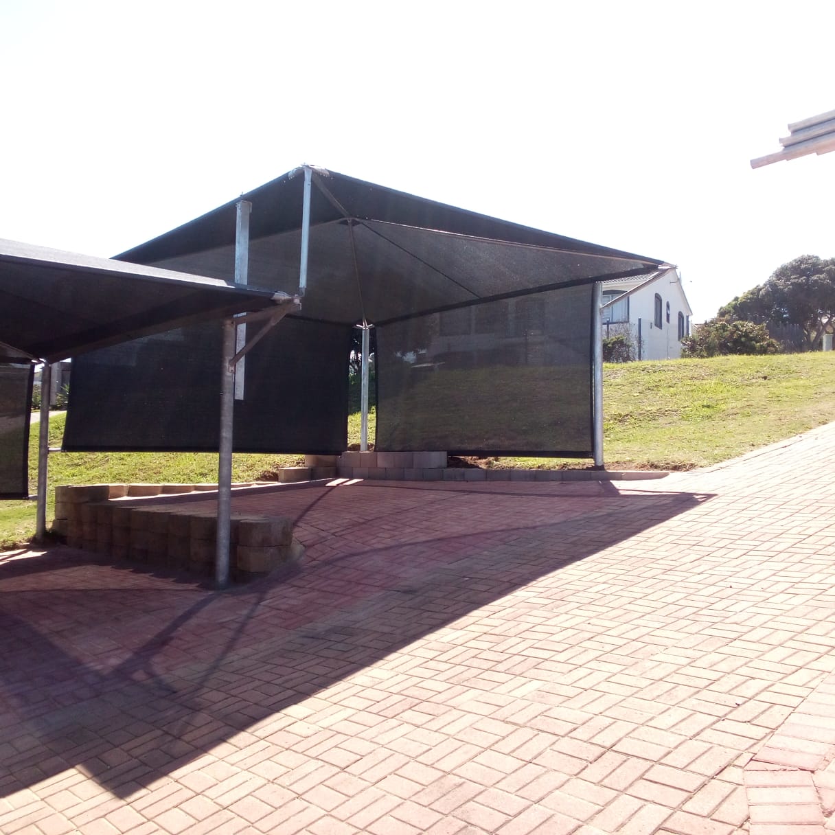 Black shade structure with metal frame on brick patio overlooking rural property and white building.