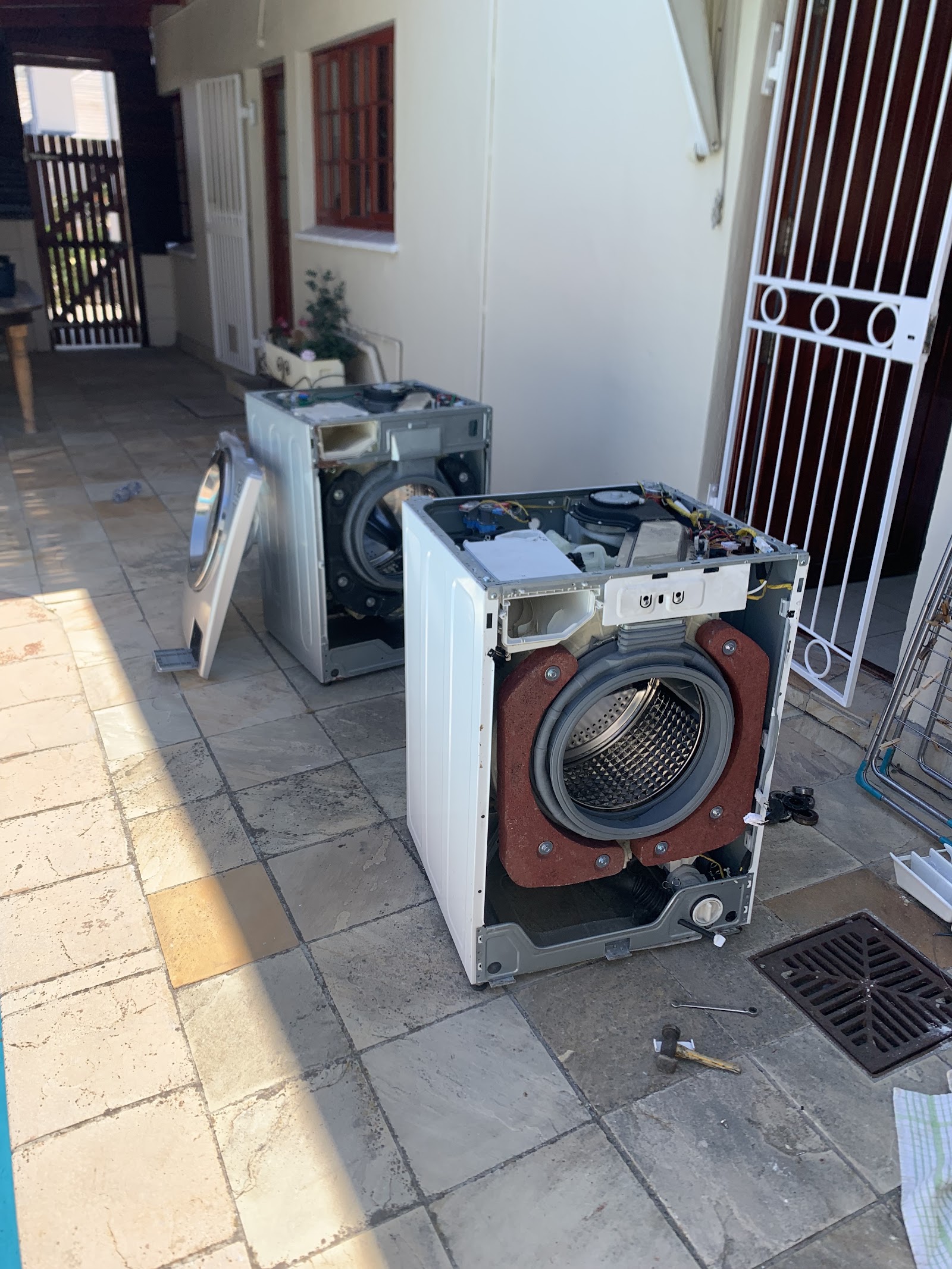 Disassembled washing machines undergoing repair work in residential courtyard with metal gates.