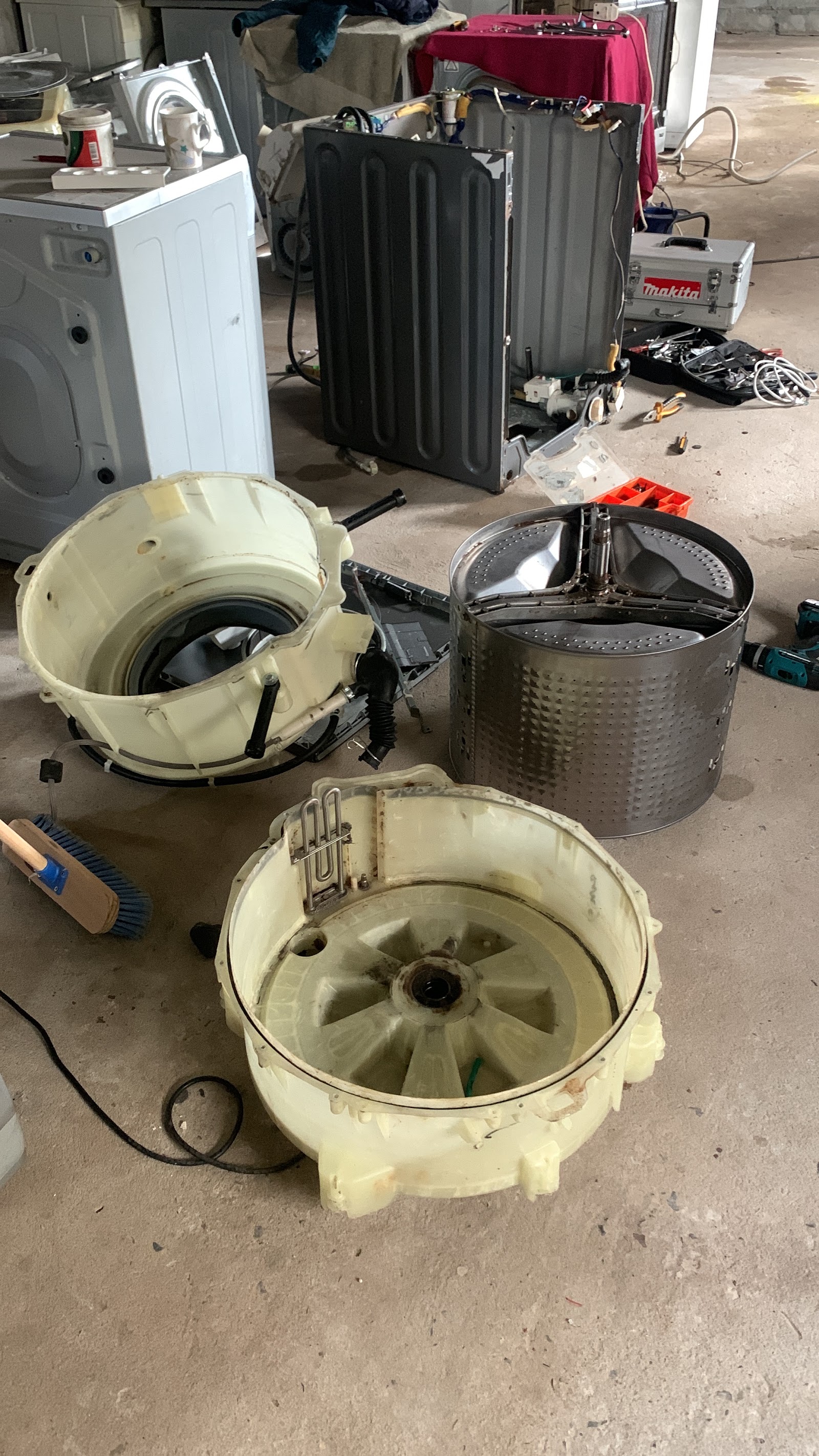 Disassembled washing machine drums, tubs, and internal components with tools on concrete work area.
