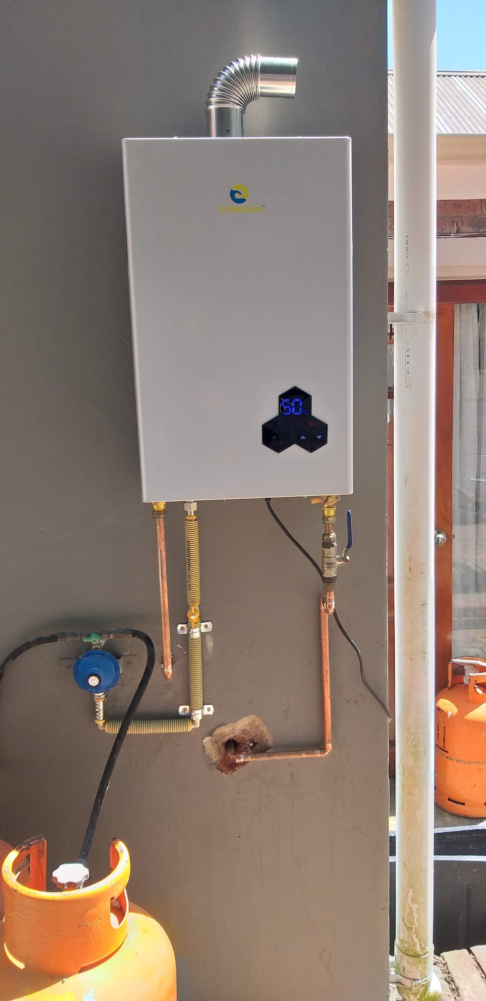 Tankless water heater with digital display showing 60°C temperature on gray wall with blue valve and gas cylinder visible.