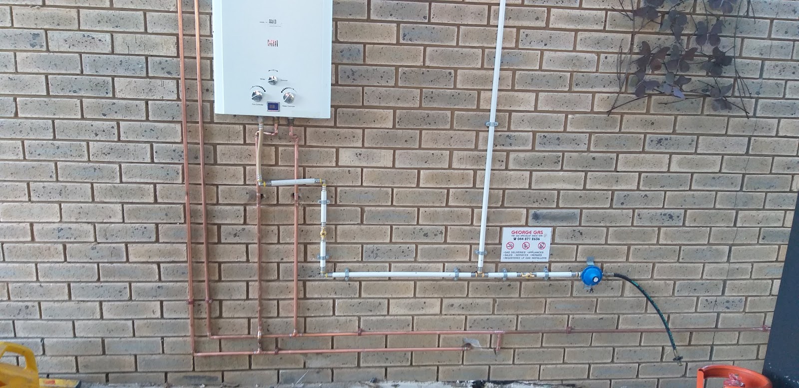 Wall-mounted gas water heater with control panel, copper and blue PVC piping, George Gas service label visible.