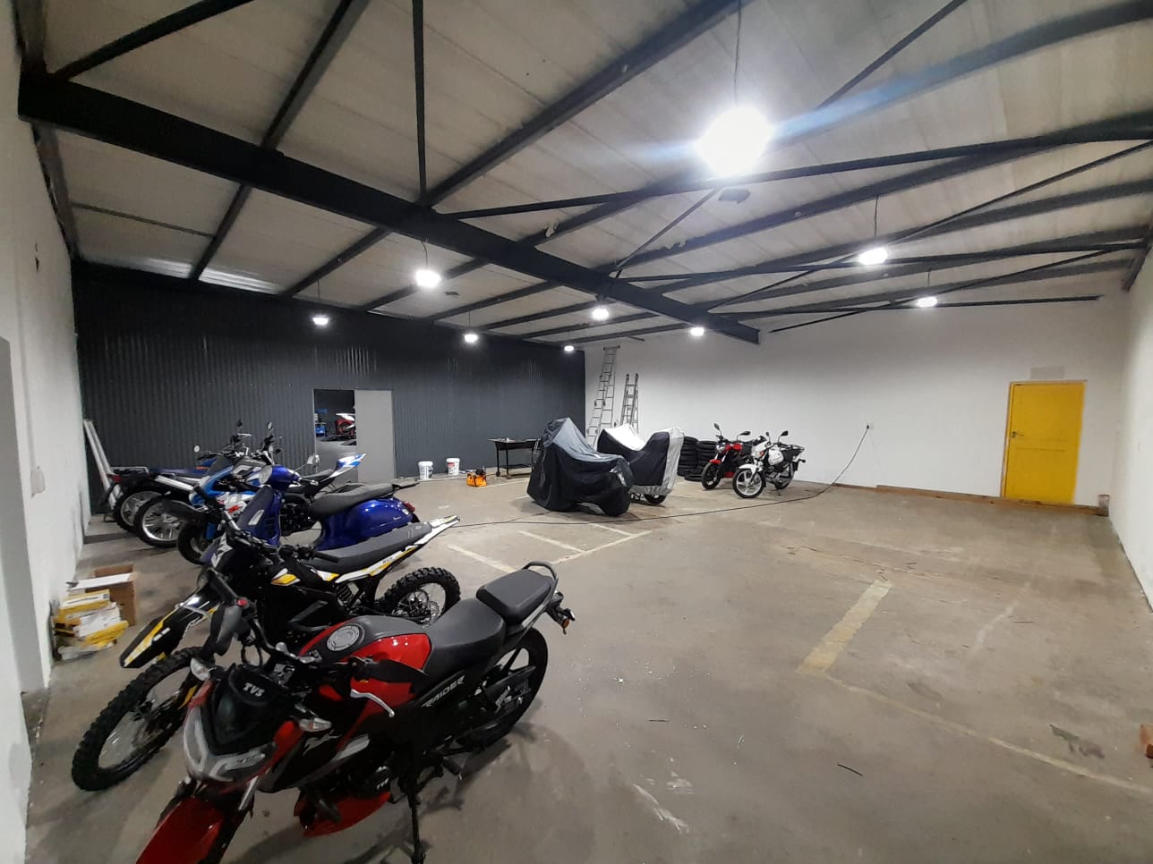 Motorcycle storage garage with red, blue motorbikes, yellow door, LED ceiling lights, concrete floor.