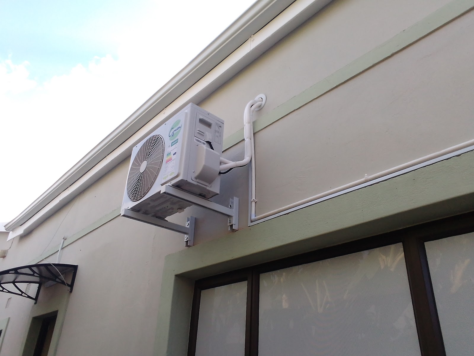 External AC compressor unit mounted on beige building wall with white refrigerant line visible.