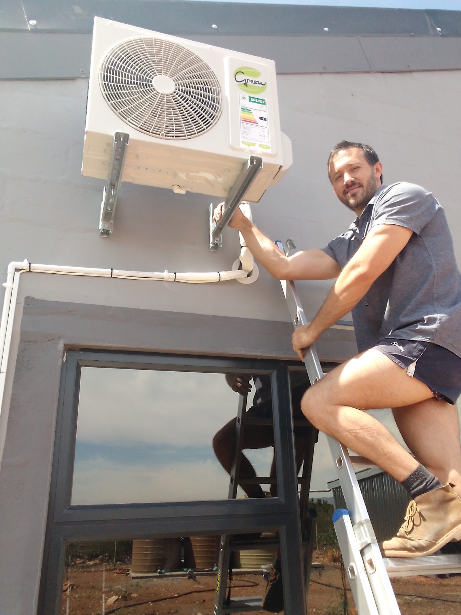 Technician on ladder installing green-branded split AC outdoor unit with energy label visible.