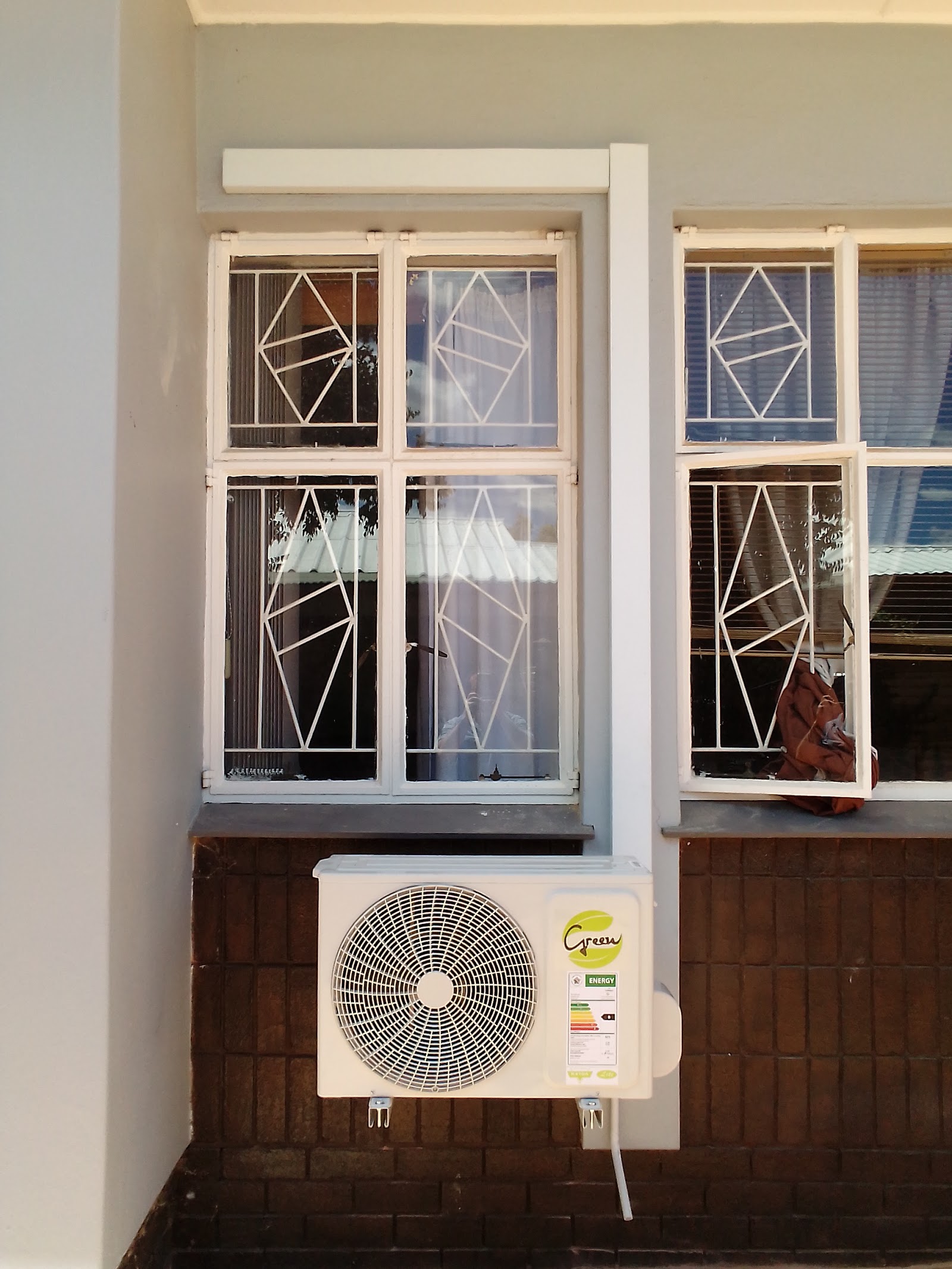 Green-branded outdoor AC compressor mounted below white-framed windows with geometric patterns.