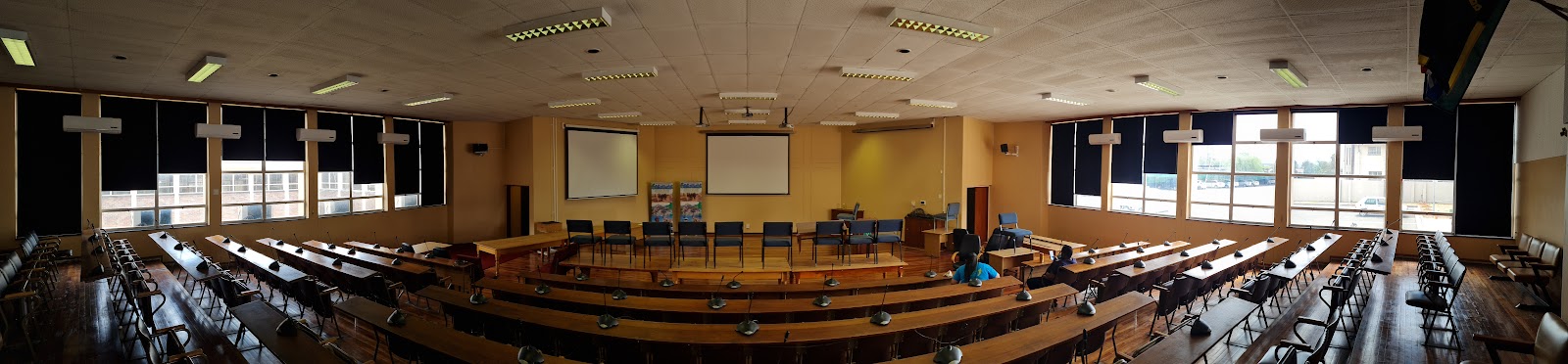 Interior of large meeting hall with curved wooden seating rows, wooden crosses, and fluorescent ceiling lights.