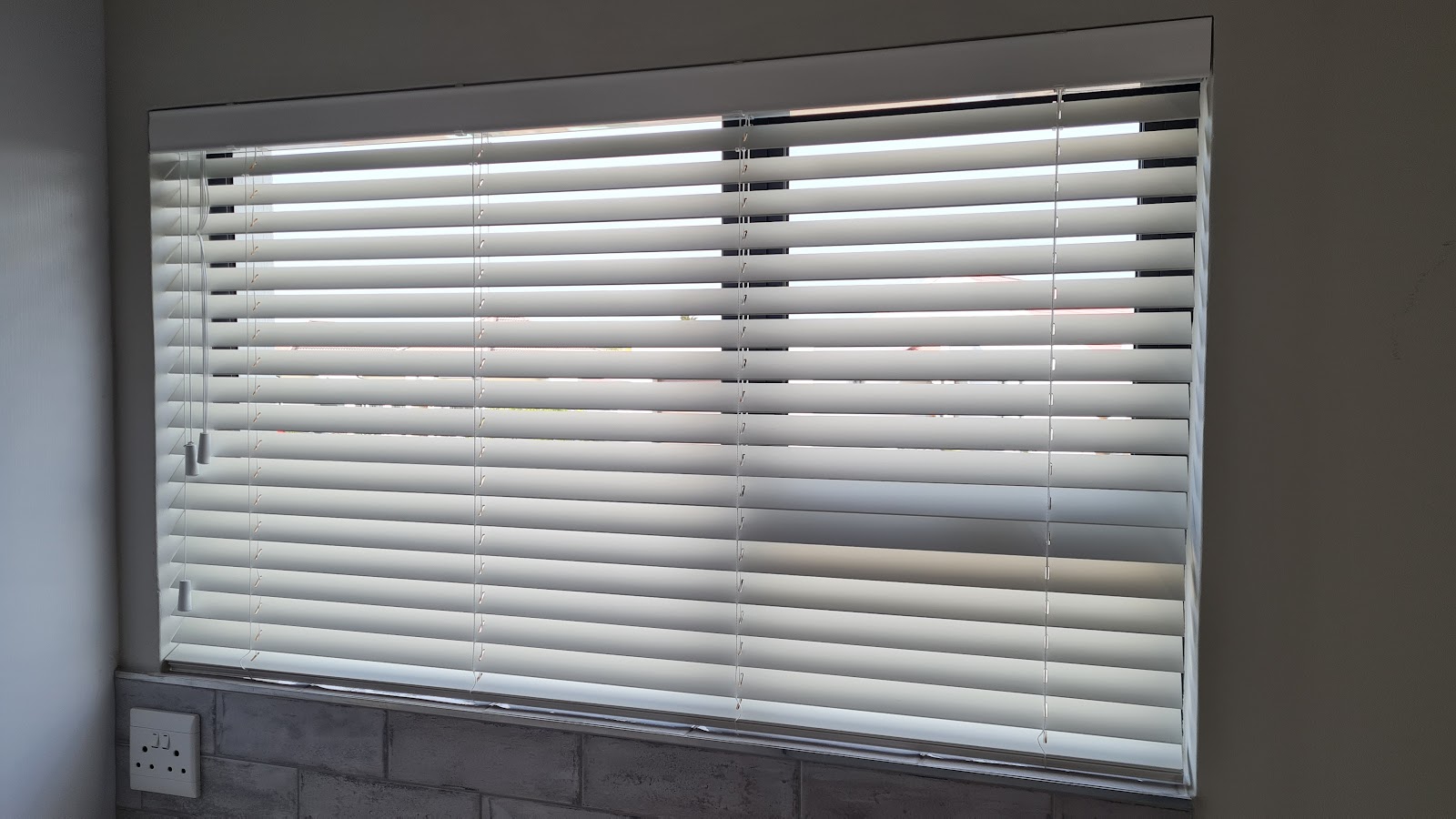 White horizontal venetian blinds installed on interior window with gray frame and electrical outlet visible below.