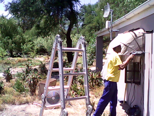 Technician in yellow shirt on ladder installing air conditioning unit at residential property.