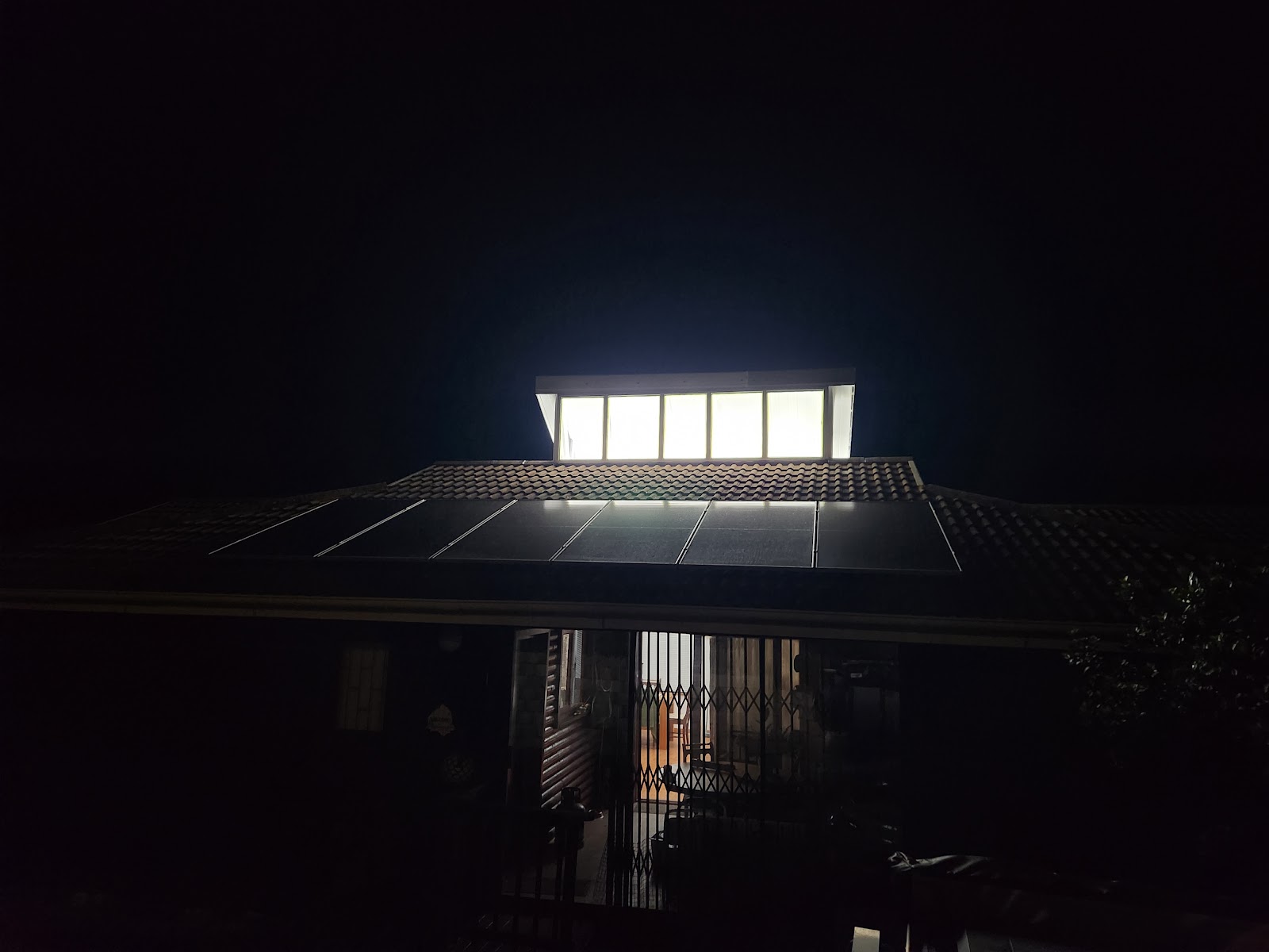 Solar panel installation on residential roof at night with illuminated skylights visible above tile roofing.