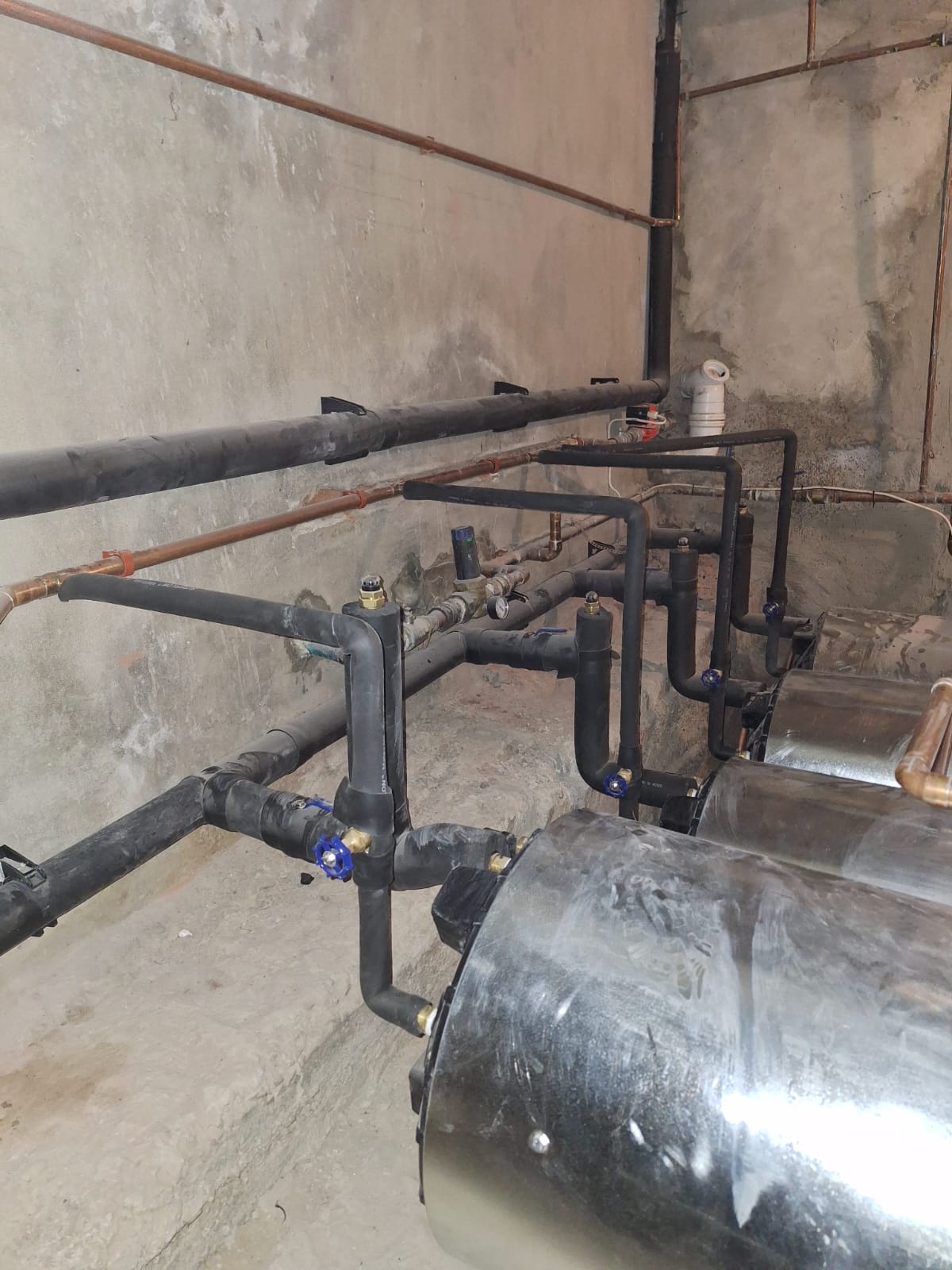 Manifold assembly with black and copper pipes, blue isolation valves, and brass fittings in concrete utility room.
