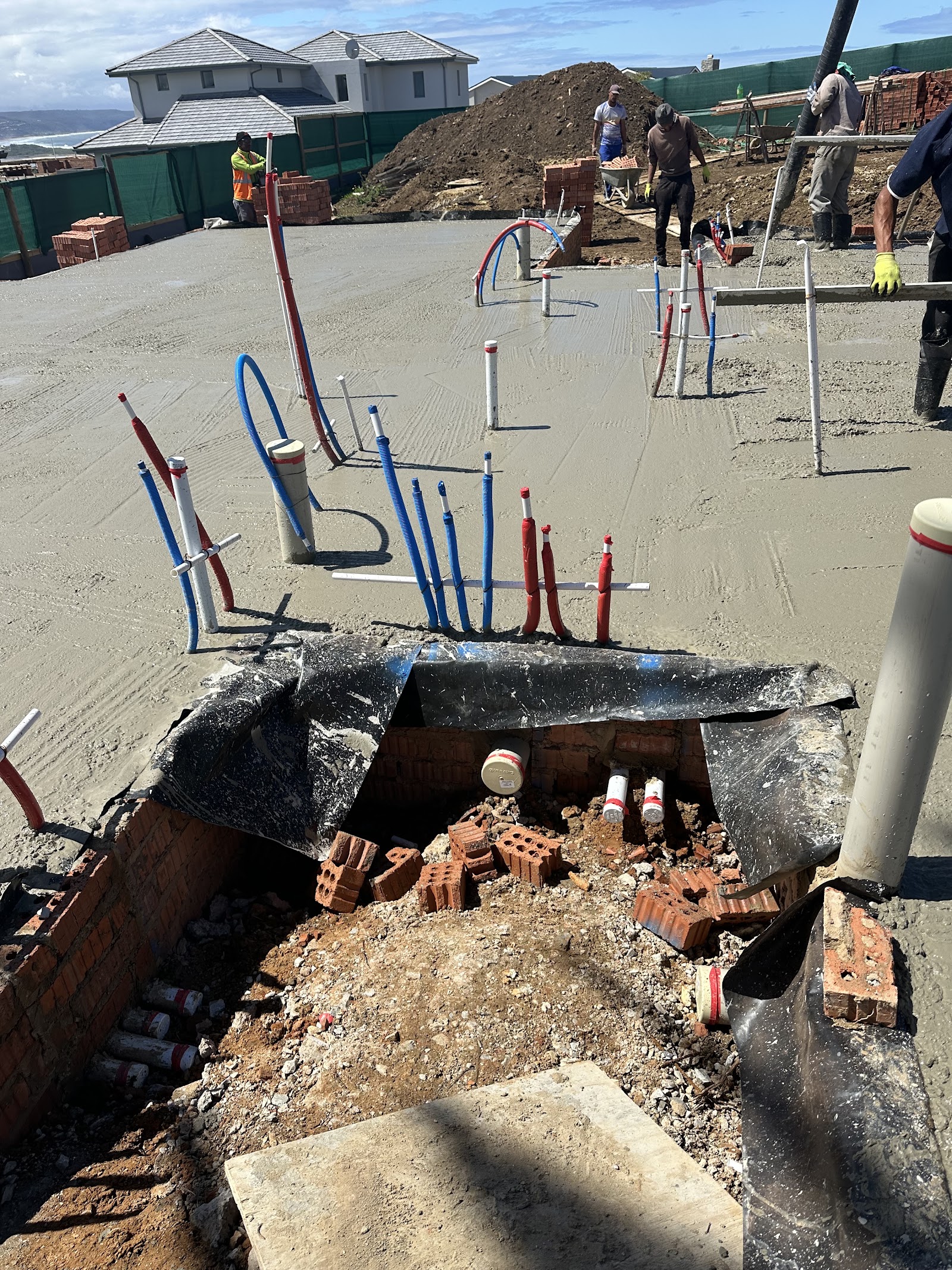 Plumbing rough-in at construction site showing blue and red pipes emerging from pit with multiple workers present.